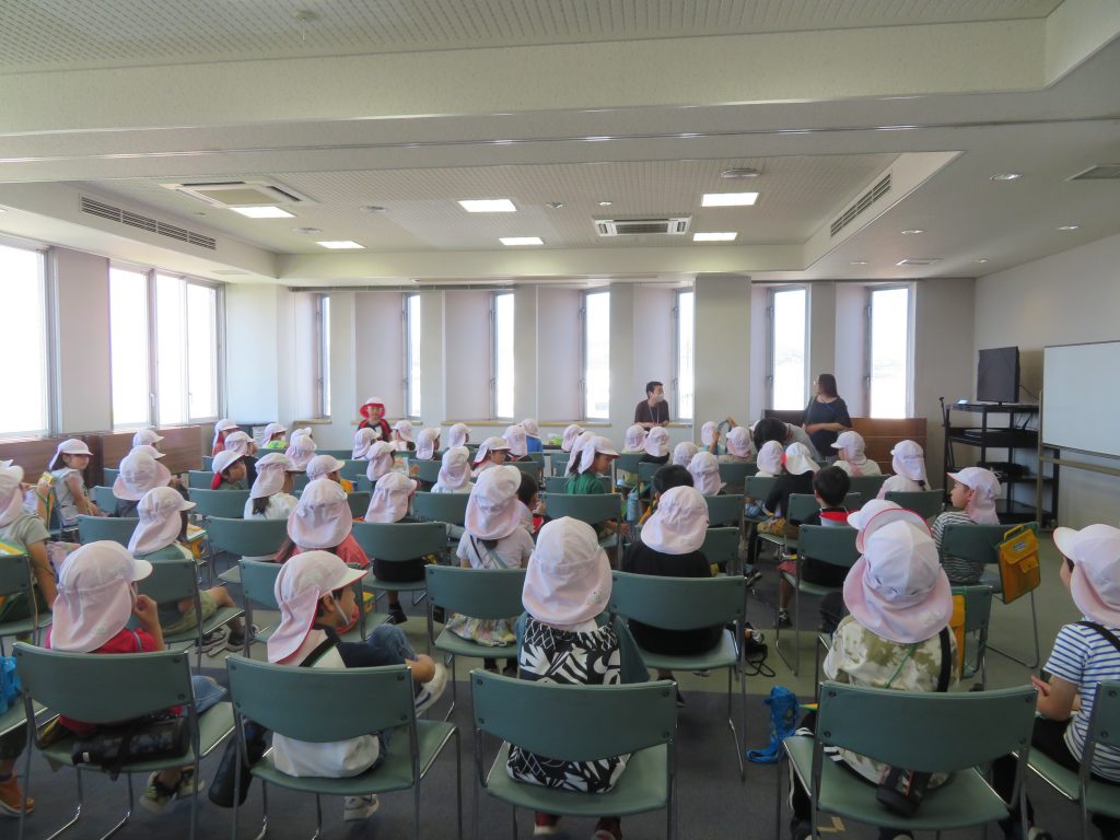 室内で帽子をかぶった多くの子どもたちが椅子に座っている様子が見える。