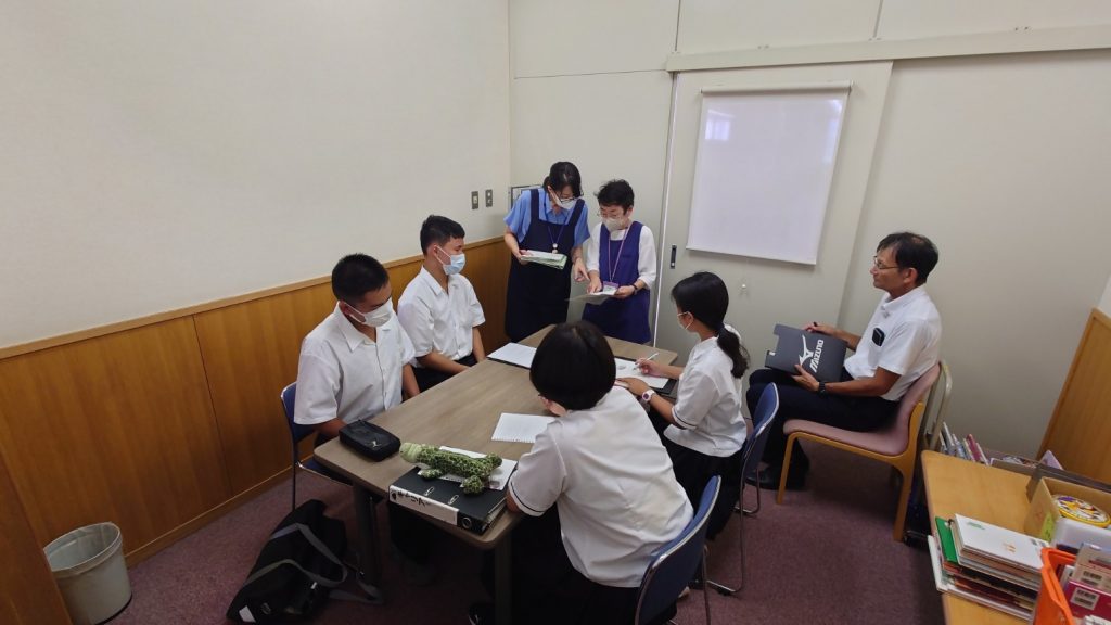 教室で数人の学生がテーブルを囲み、誰かが立って話している様子の画像