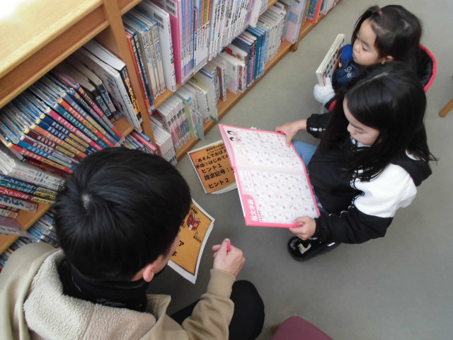 図書館で本を見ながら話し合う三人の学生がいる様子。