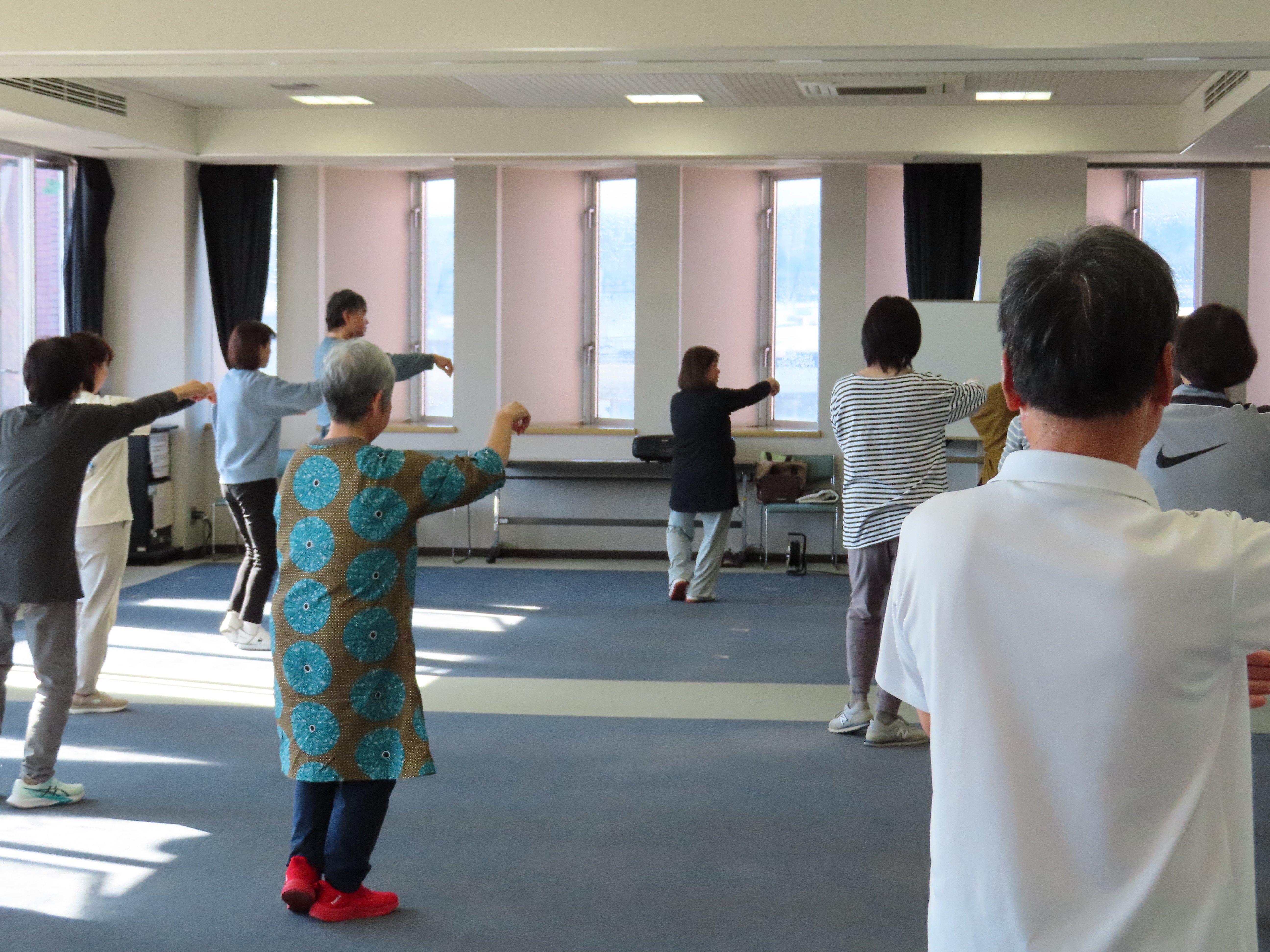 数人の人々が広い室内で前に腕を伸ばす太極拳の動作をする参加者