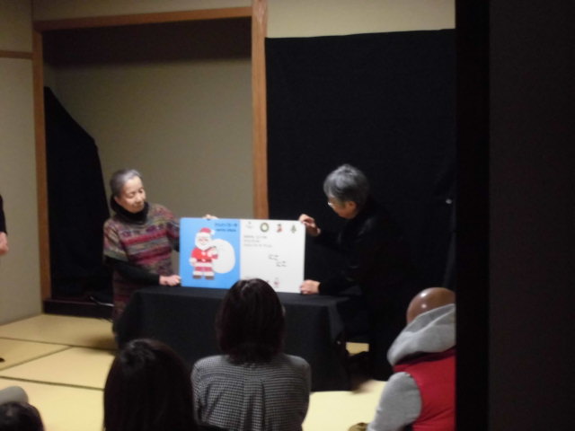 二人の人が絵本を持っている様子が映っているシーンで、観客が前方で見守っている。