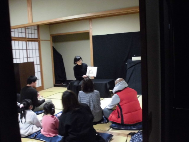 子供たちが集まっている部屋で、読み聞かせをしている人が本を持っている様子。