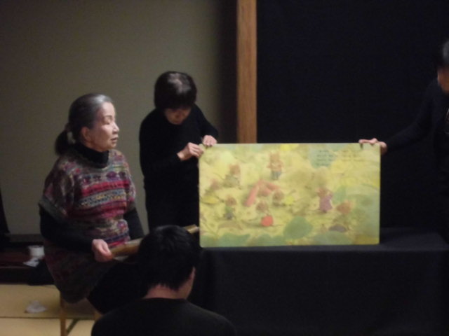 二人の人物が色とりどりの絵を掲げているシーンで、手前には座っている人物がいる。