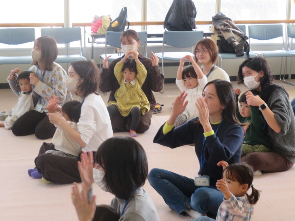 多くの大人と子供が座って手を挙げている室内の様子。