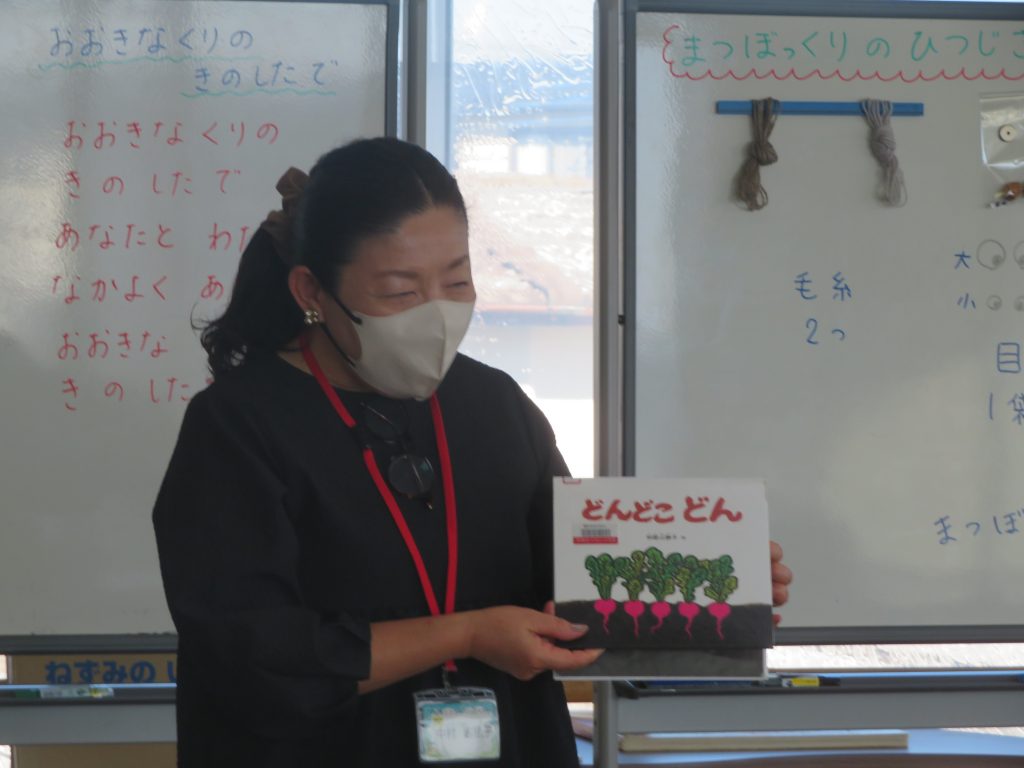 ホワイトボードの前で絵本を持つ人物が写っている画像。