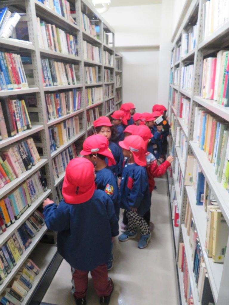 図書館の棚の間で本を見ている赤い帽子をかぶった子どもたちの集団。