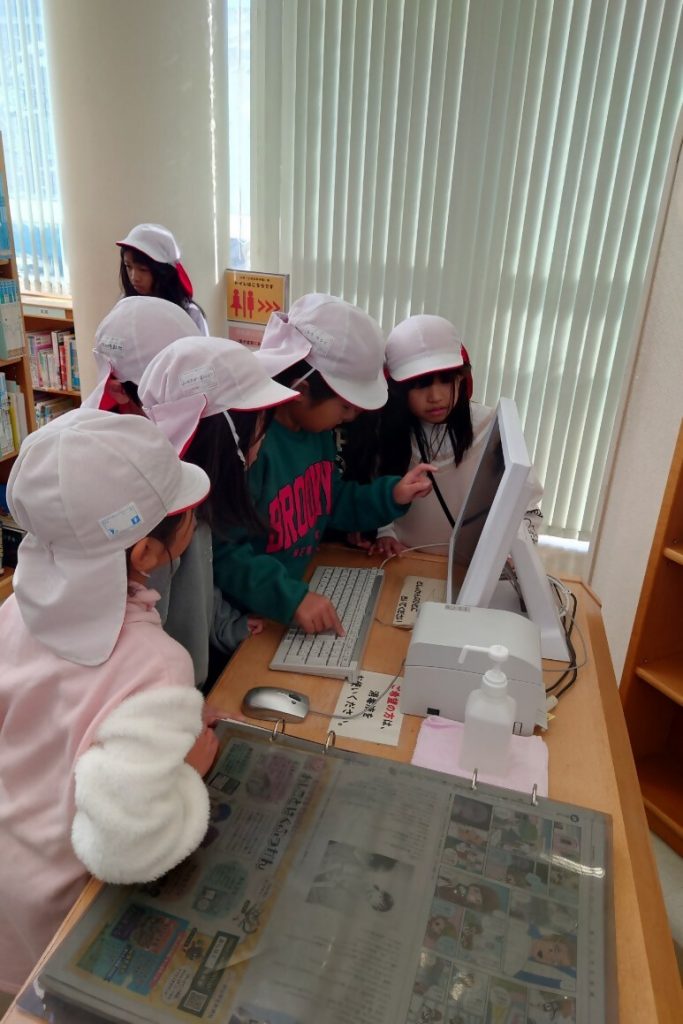 白い帽子をかぶった子どもたちがコンピュータの前で作業している図書室の内部の様子。