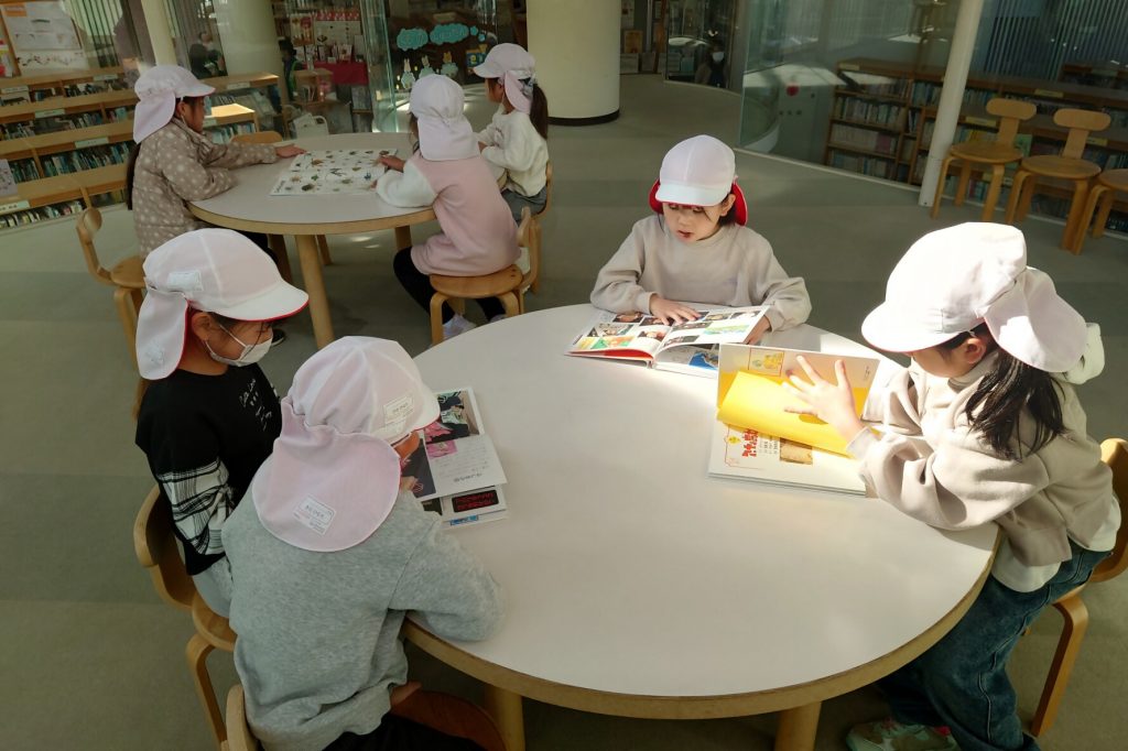 幼い子供たちが白い帽子をかぶりながらテーブルで本を読んでいる様子。