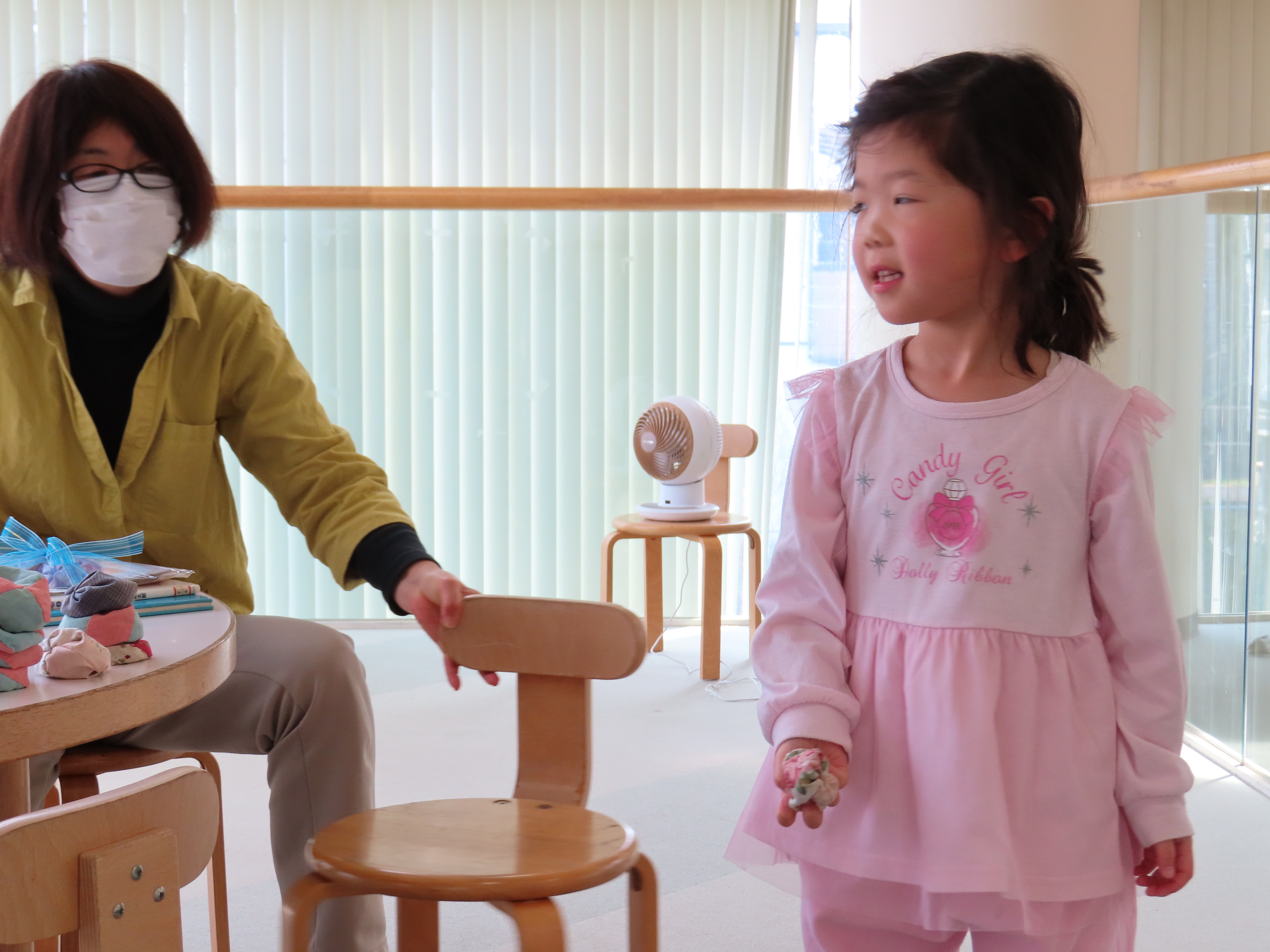 テーブルのそばにマスクをした大人が座って椅子に手を伸ばしもう一方でピンクの服を着た子どもが立ってお手玉を手に持っている様子