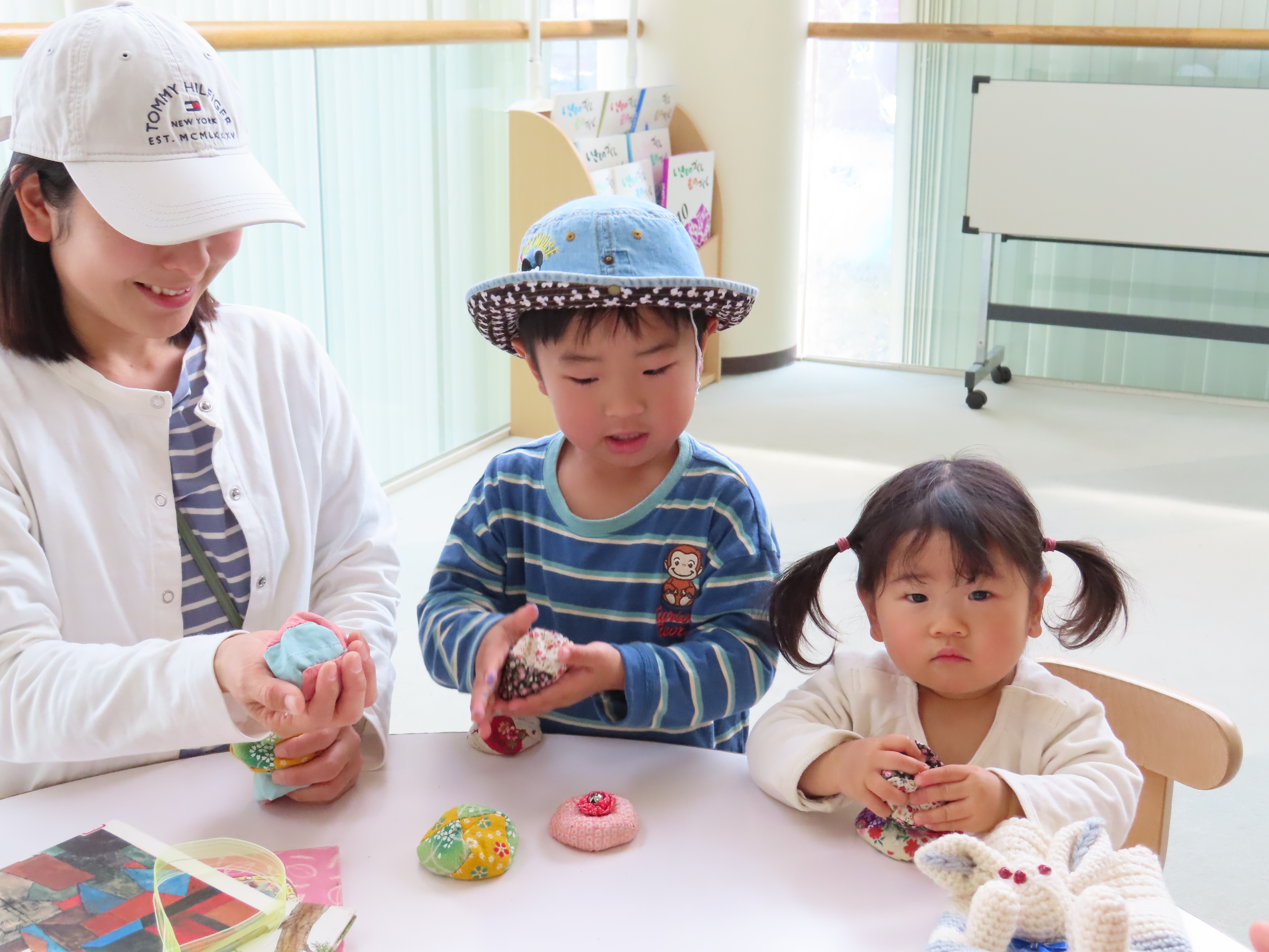 室内のテーブルでお手玉を手に取る帽子の男児と左右に座る大人と髪をツインテールにした女児