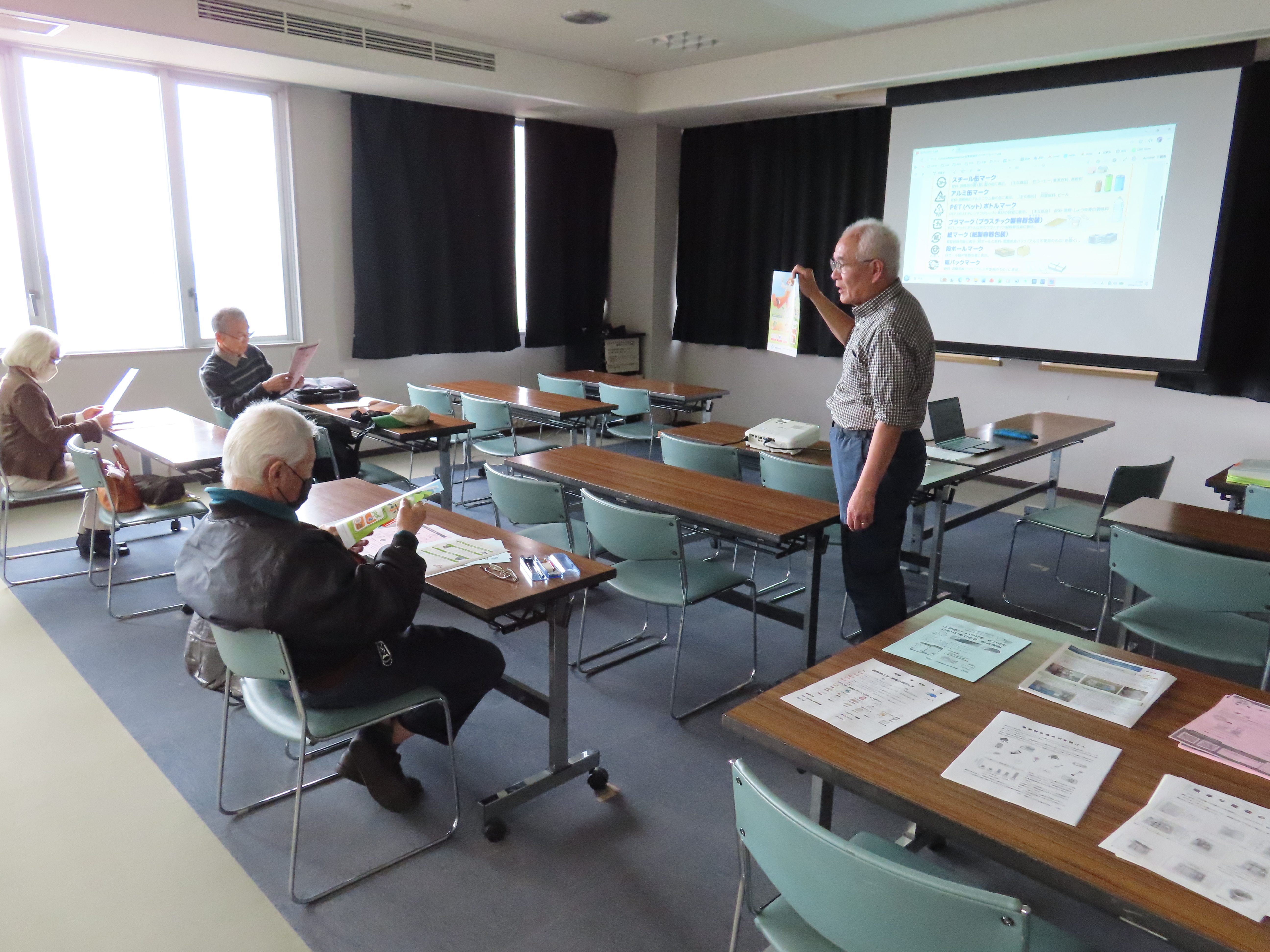 教室の前でスクリーンに投影しながら資料を手にして立つ先生と席で資料を手にする数人の参加者がいる風景