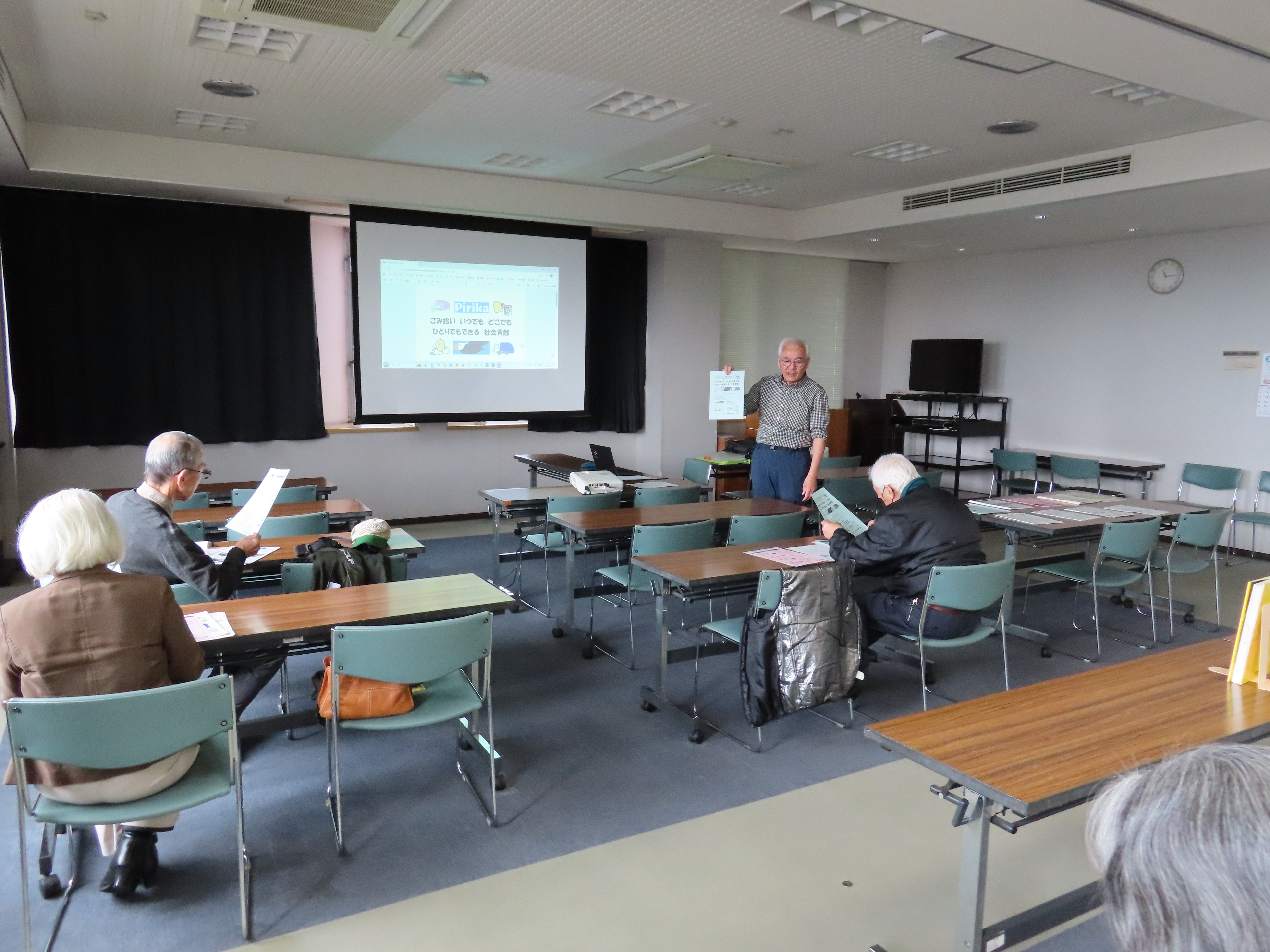 教室のような部屋でスクリーンに投影された資料を前に数人の参加者が席に座って資料を読み先生が前に立っている