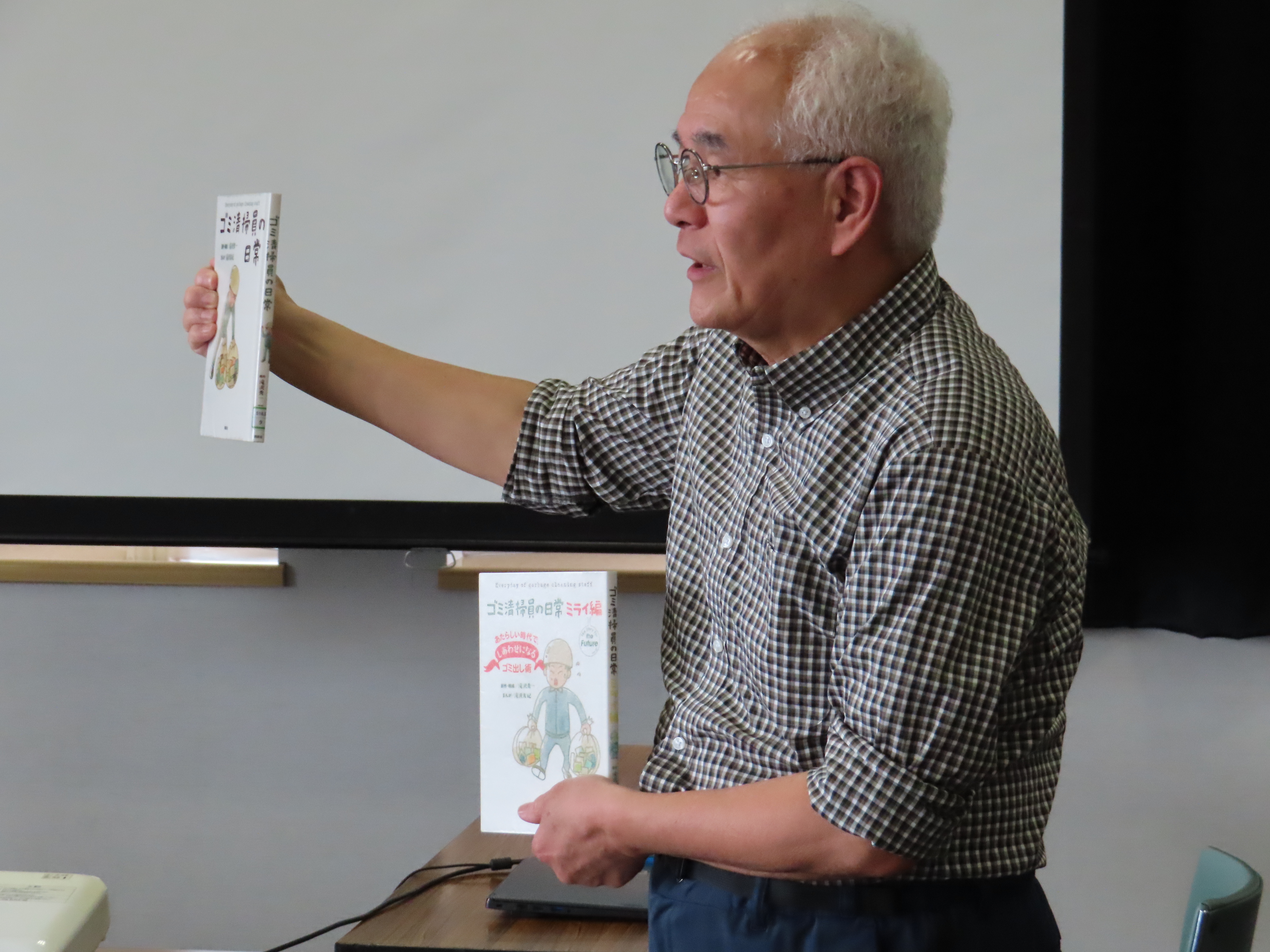 先生が教室の前で一冊を高く掲げもう一冊を下に持って見せている