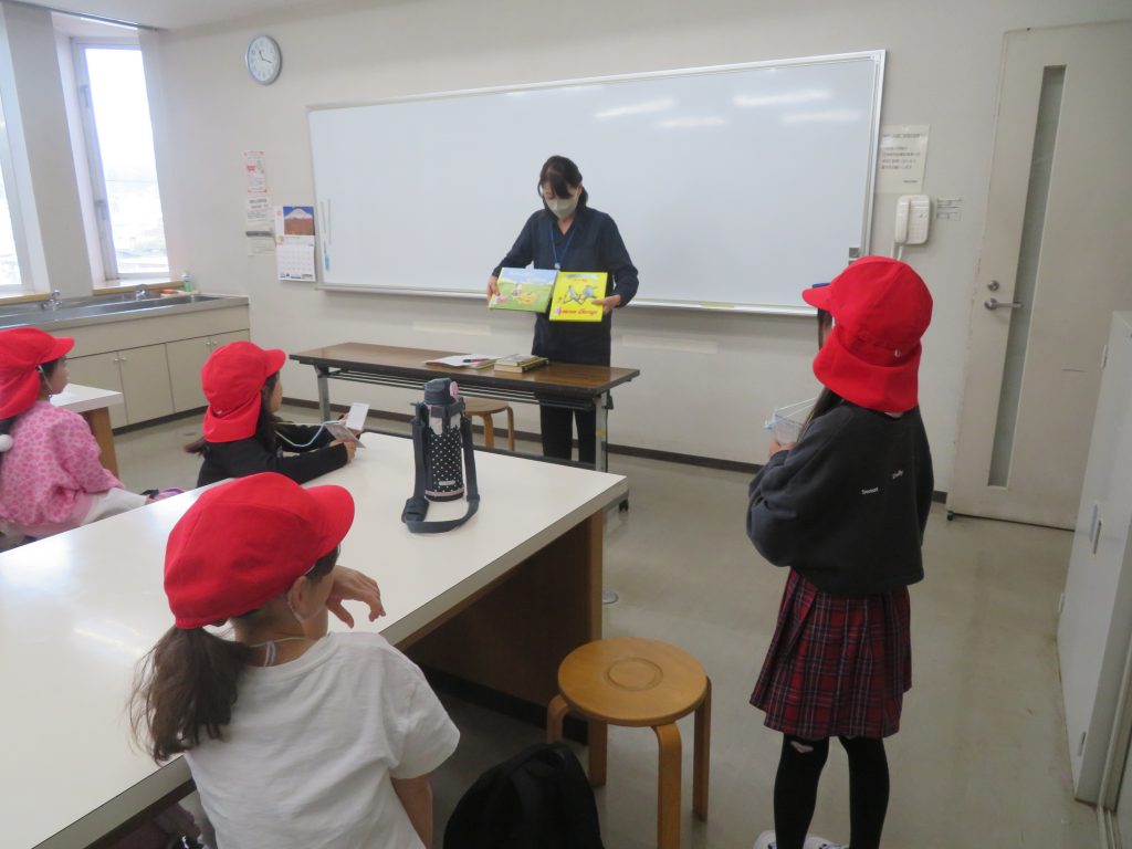 教室で赤い帽子をかぶった子供たちが教師の話を聞いている様子。