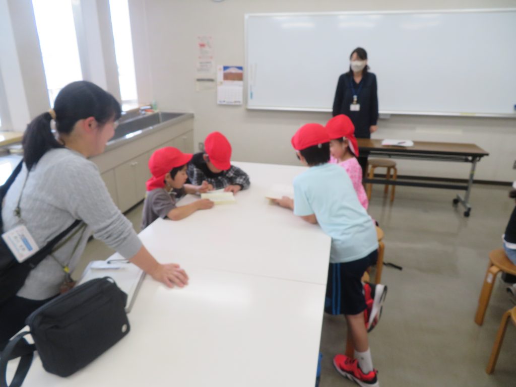 教室で赤い帽子をかぶった子供たちがテーブルに集まり、作業をしている様子が写っている画像。