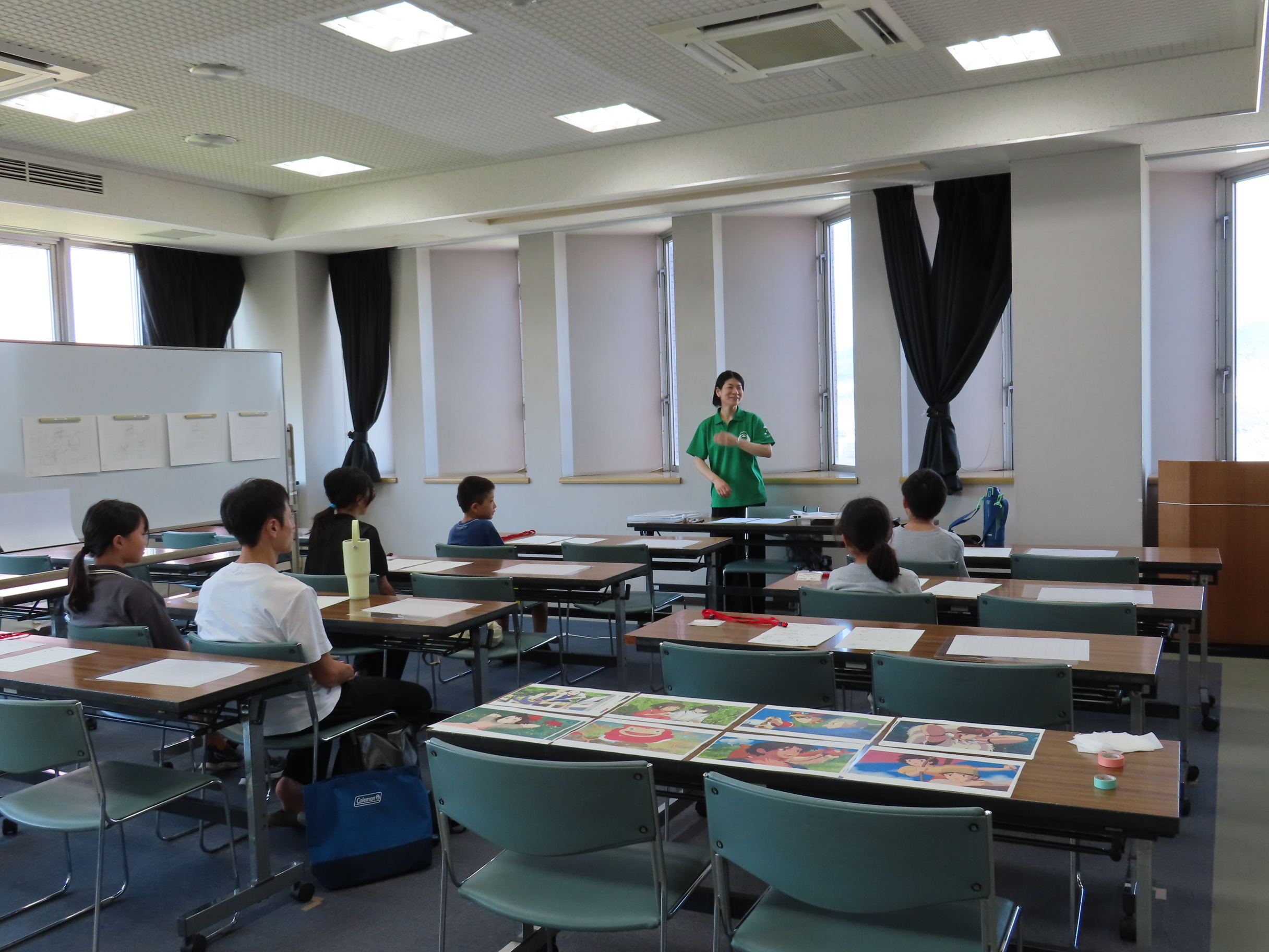 教室で机に座る子どもたちが前方で立つ指導者の話を聞きながら教材や作品が並ぶ机を見ている風景