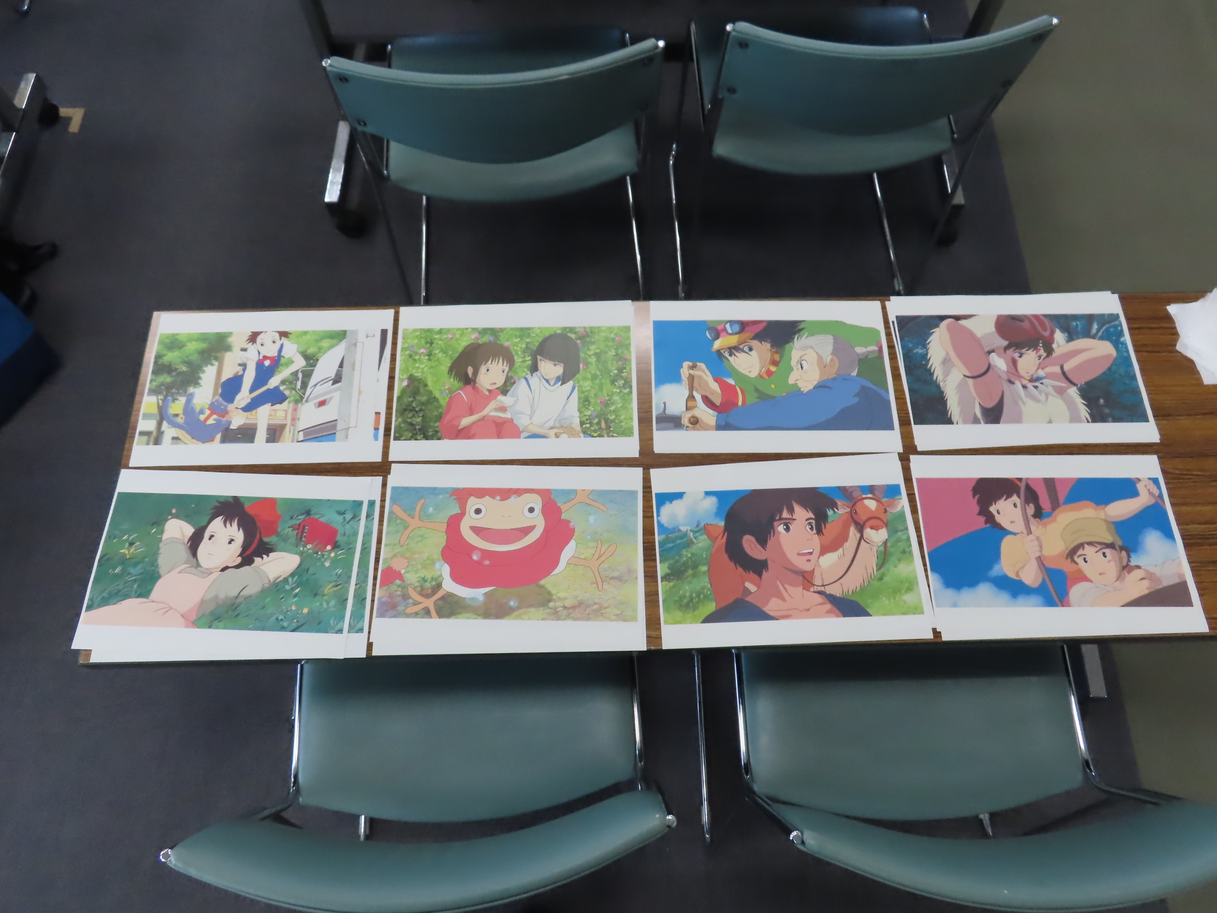 会議用の椅子が並ぶテーブルの上に色とりどりのアニメイラストが八枚整列して並べられている風景