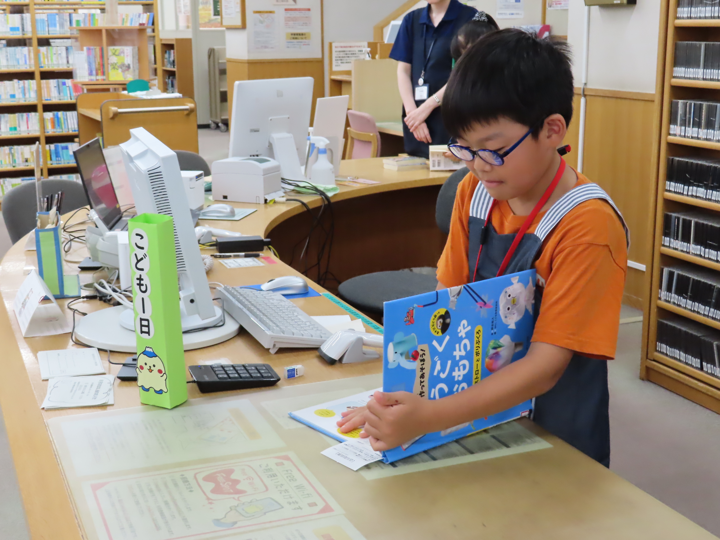 図書館のカウンターで眼鏡をかけた子どもが青い絵本を手に貸出手続きをしている様子