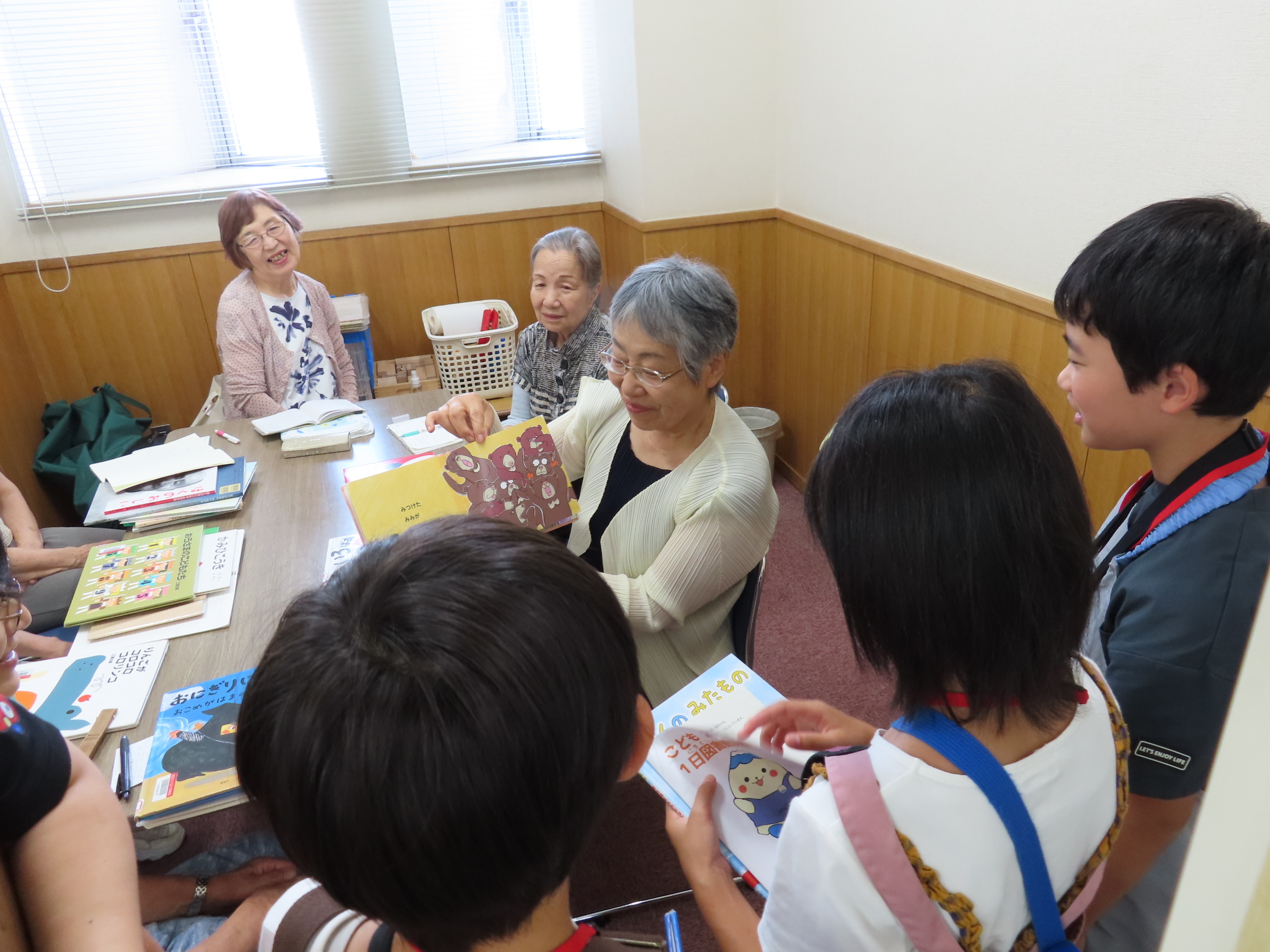 高齢の女性たちと子どもたちがテーブルを囲んで本を開きながら和やかに交流している場面