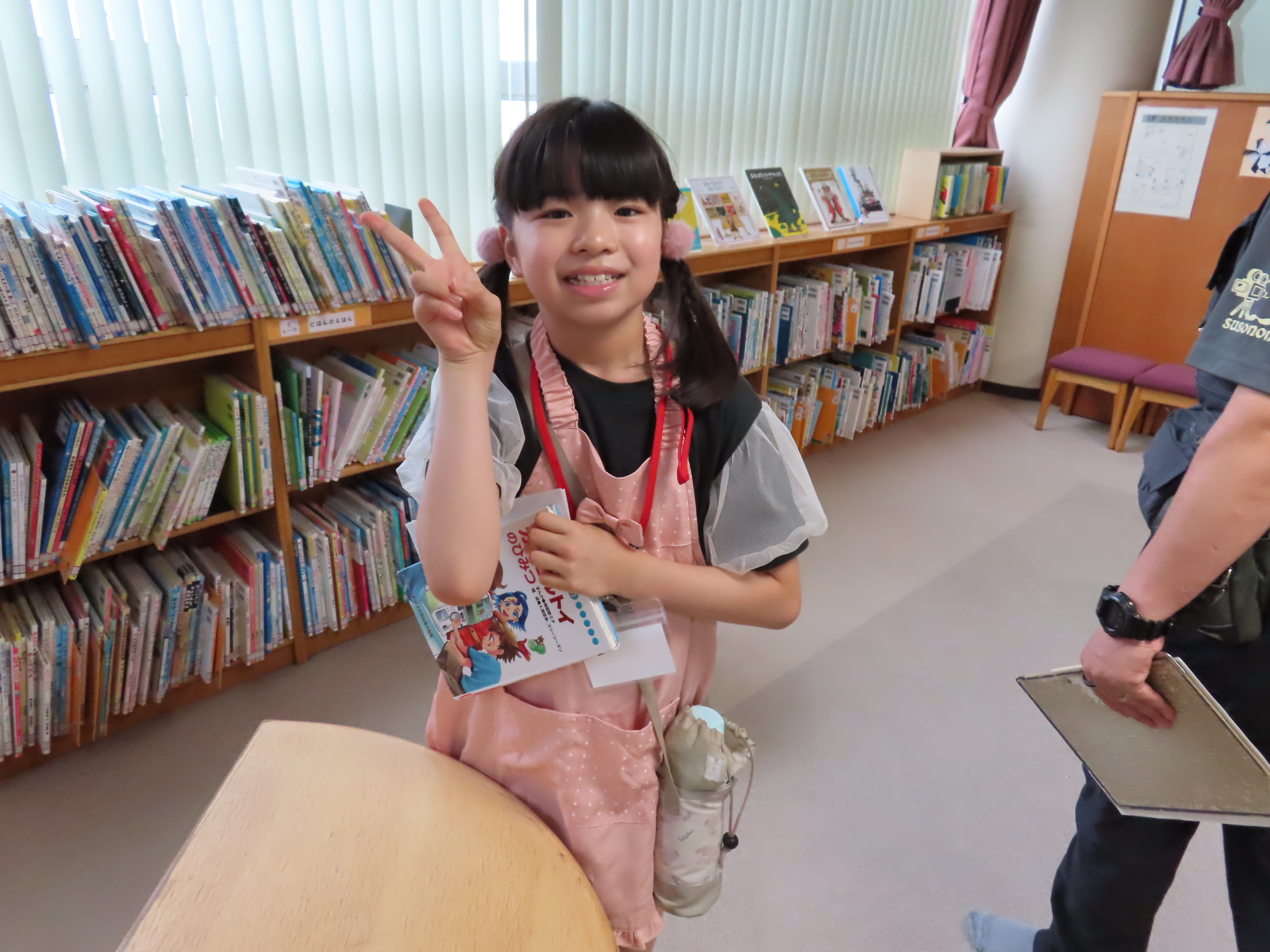 図書館の本棚の前でピースサインをしている子どもがピンクのエプロンを着て本を持っている