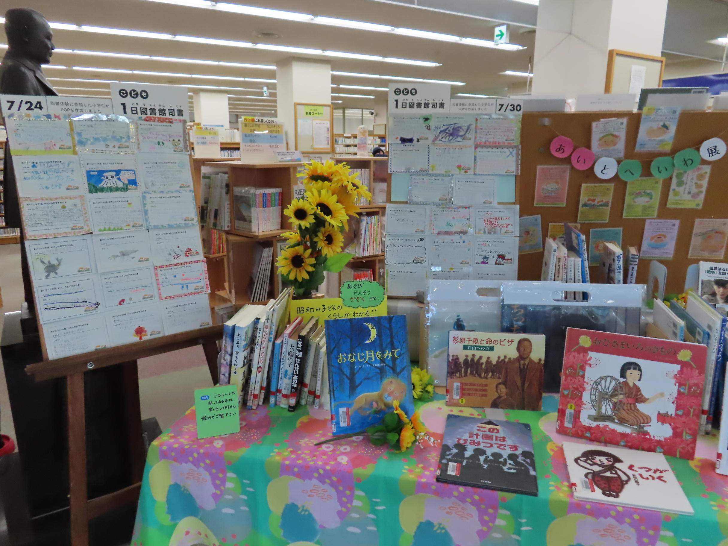 図書館の棚の前にひまわりが飾られ子ども向けの絵本がテーブルに並べられポップが背景に立てかけられた展示