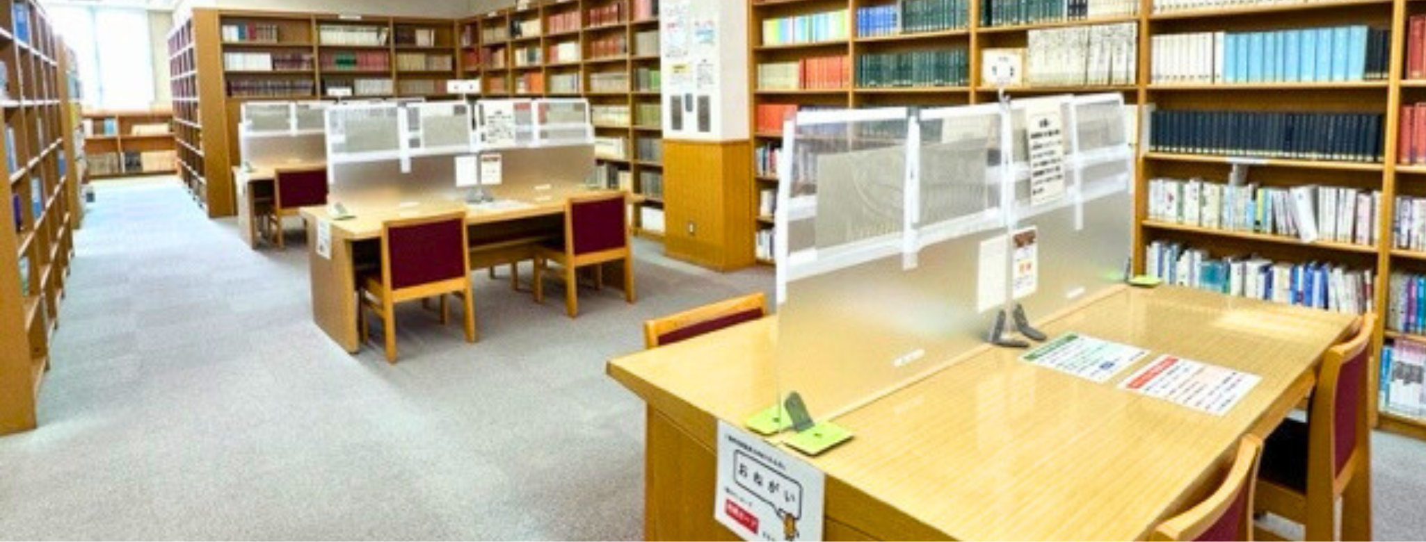 図書館の閲覧スペースで本棚に囲まれた机と椅子に透明な仕切り板が並んでいる