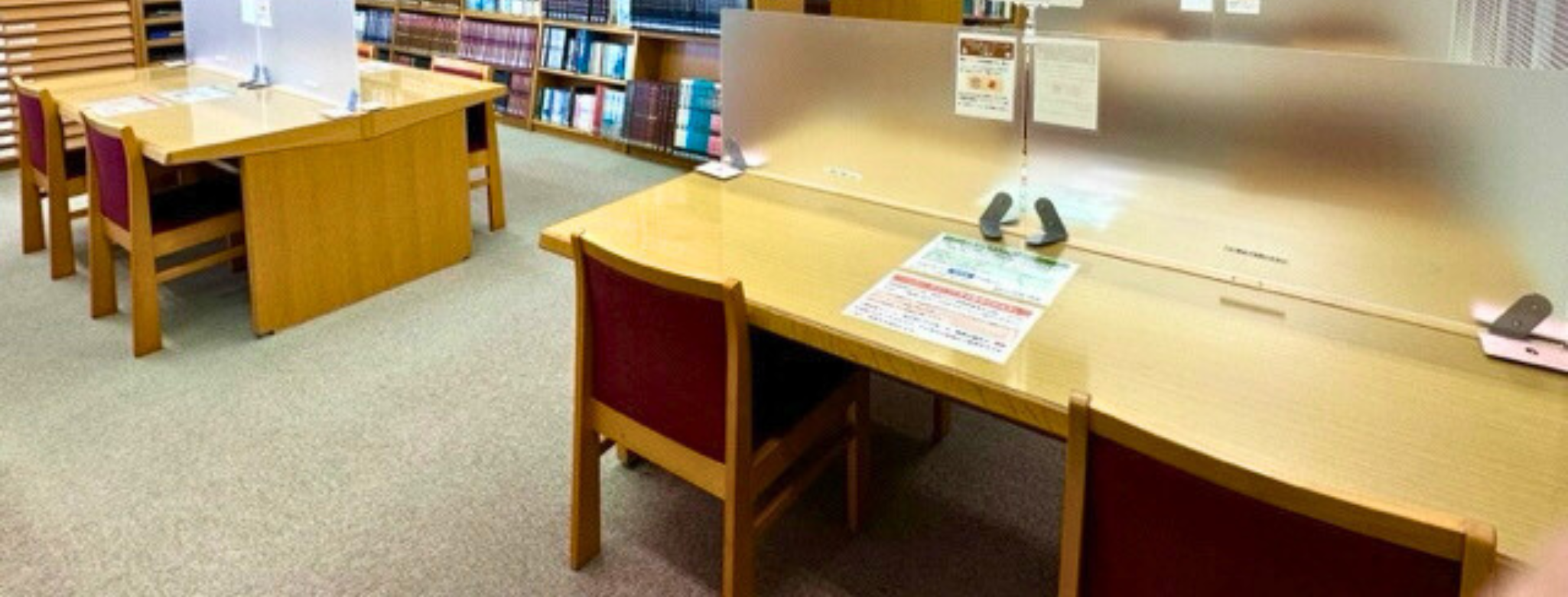 木製の学習机と椅子が並び背後に本棚がある図書館内スペース