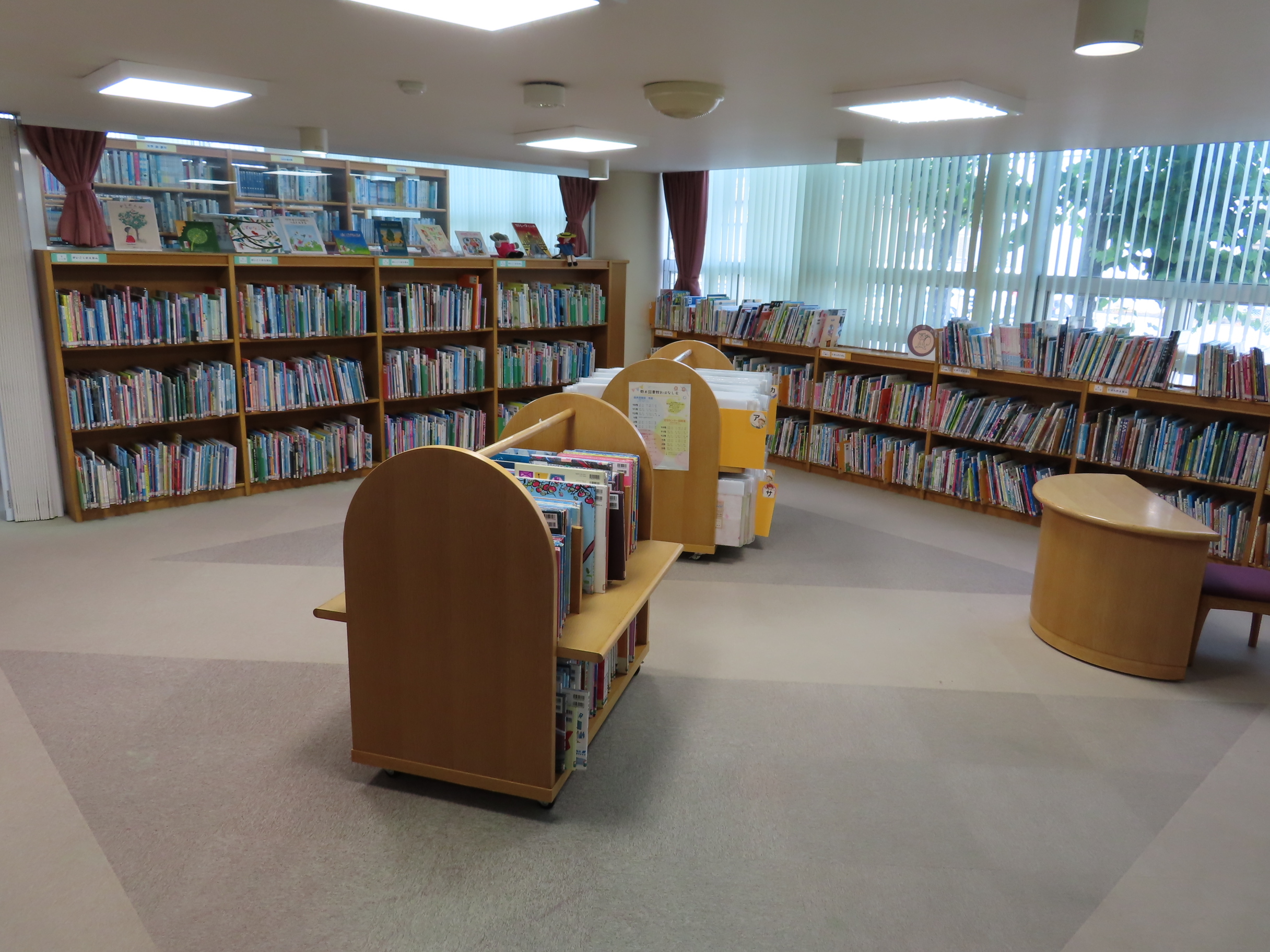 明るい窓から自然光が入る低い本棚と小さなベンチや丸いテーブルが並んだ児童向けの図書コーナー