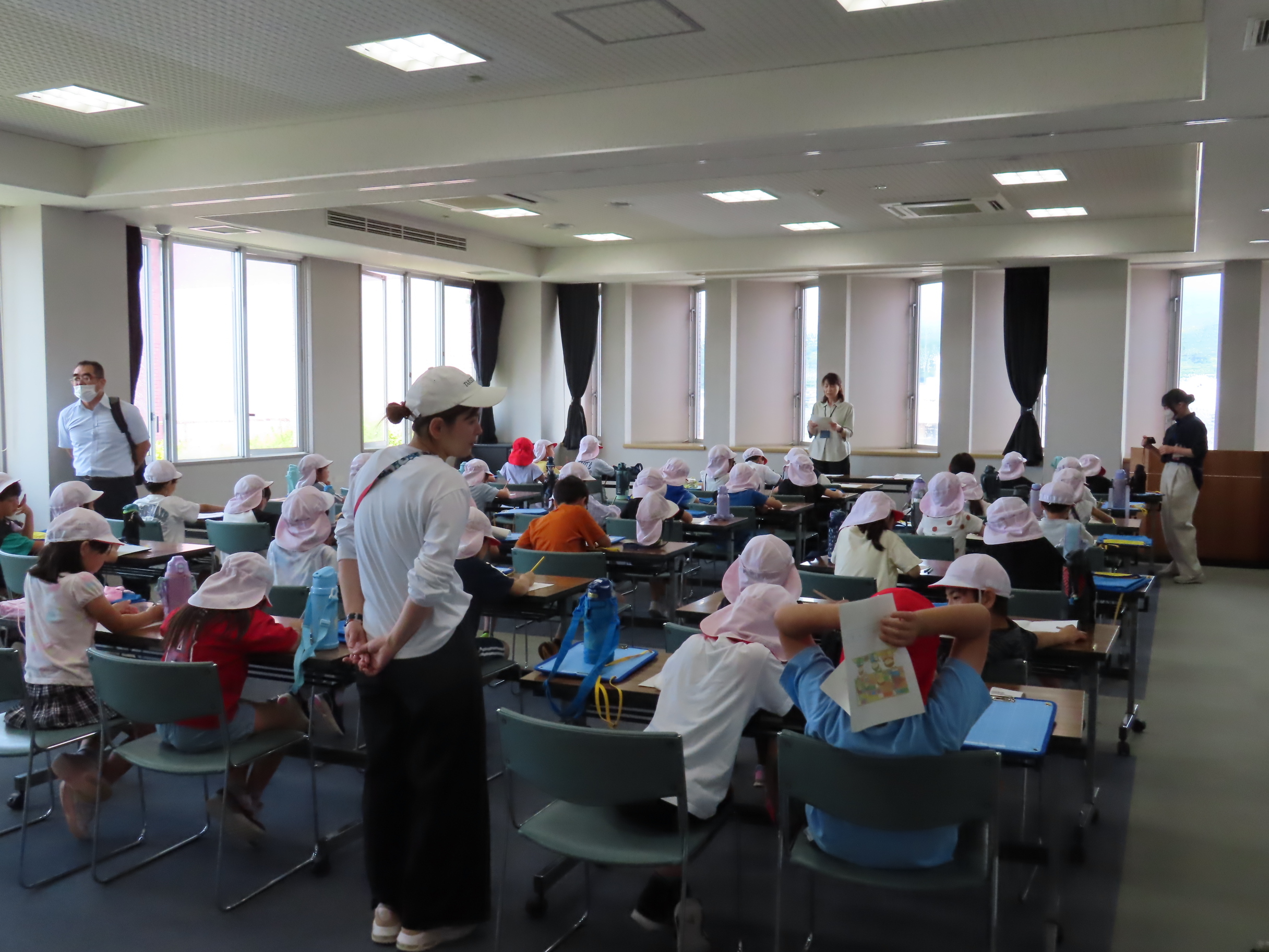 白い帽子をかぶった子どもたちが机に向かい先生が教室内を歩き回る学習の様子