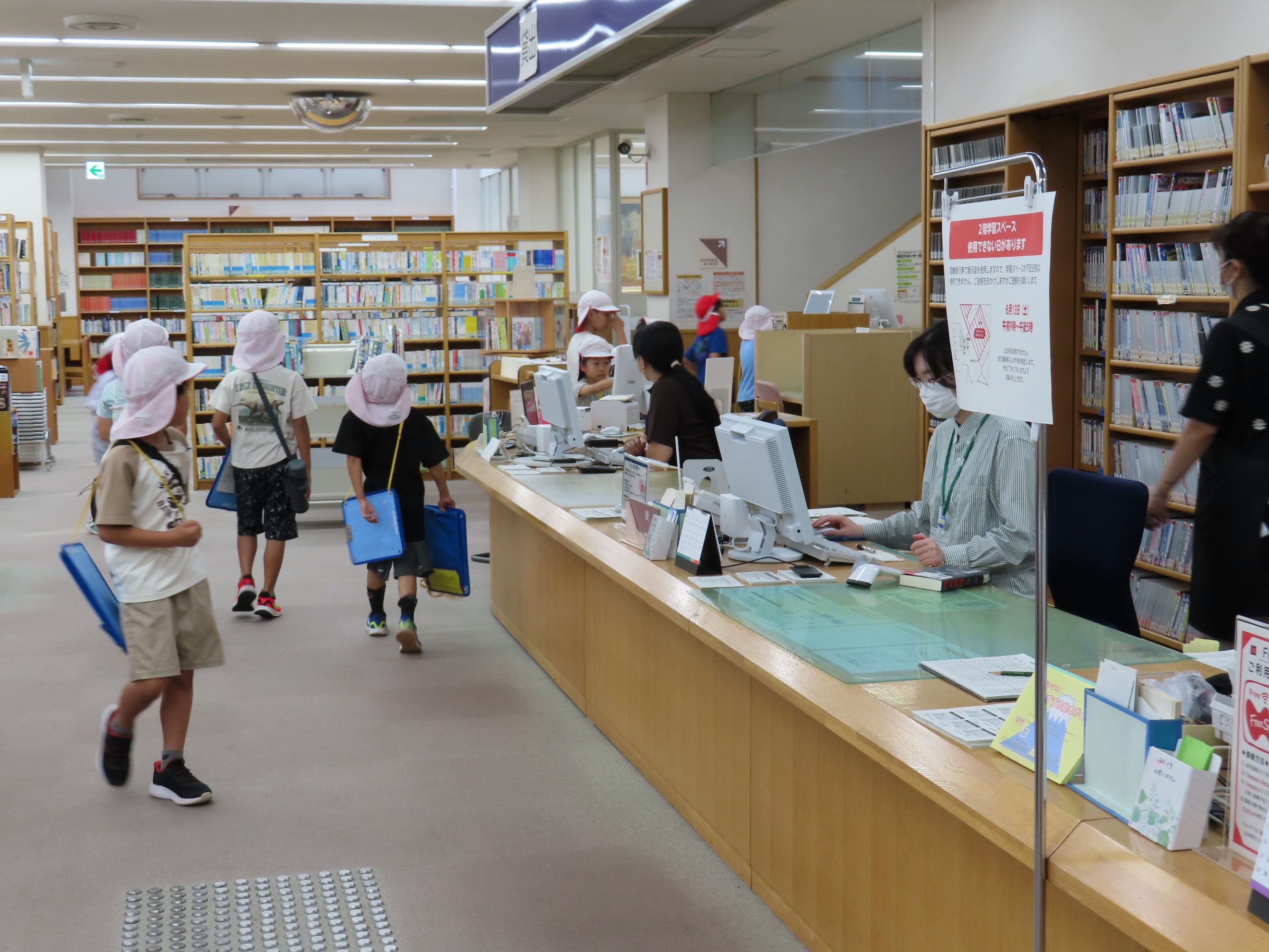 図書館の閲覧室で帽子をかぶった子どもたちがカウンターに並び職員がコンピューターで対応している様子