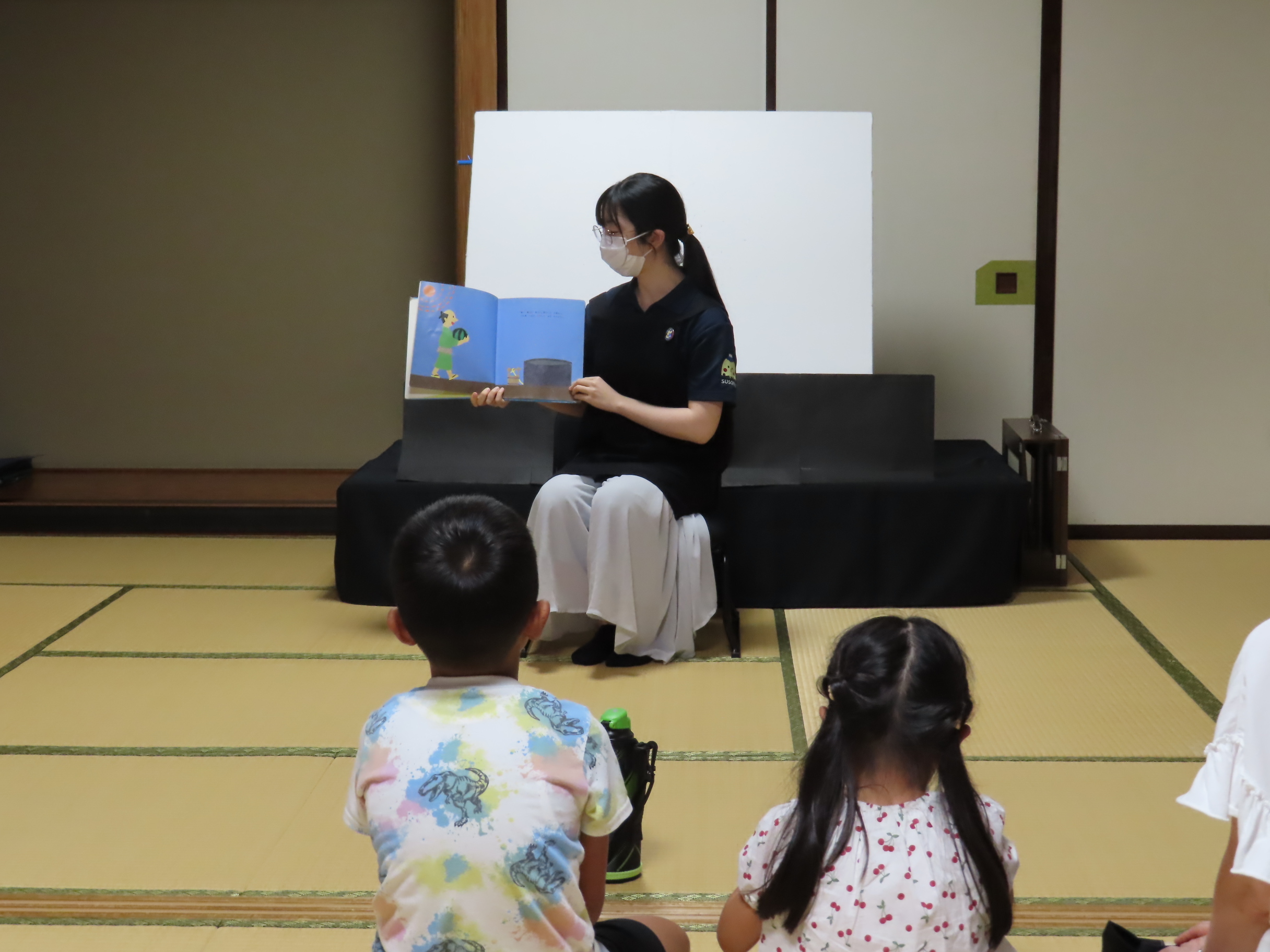 畳の上に座った子どもたちを前に女性が絵本を見せながら読み聞かせしている場面