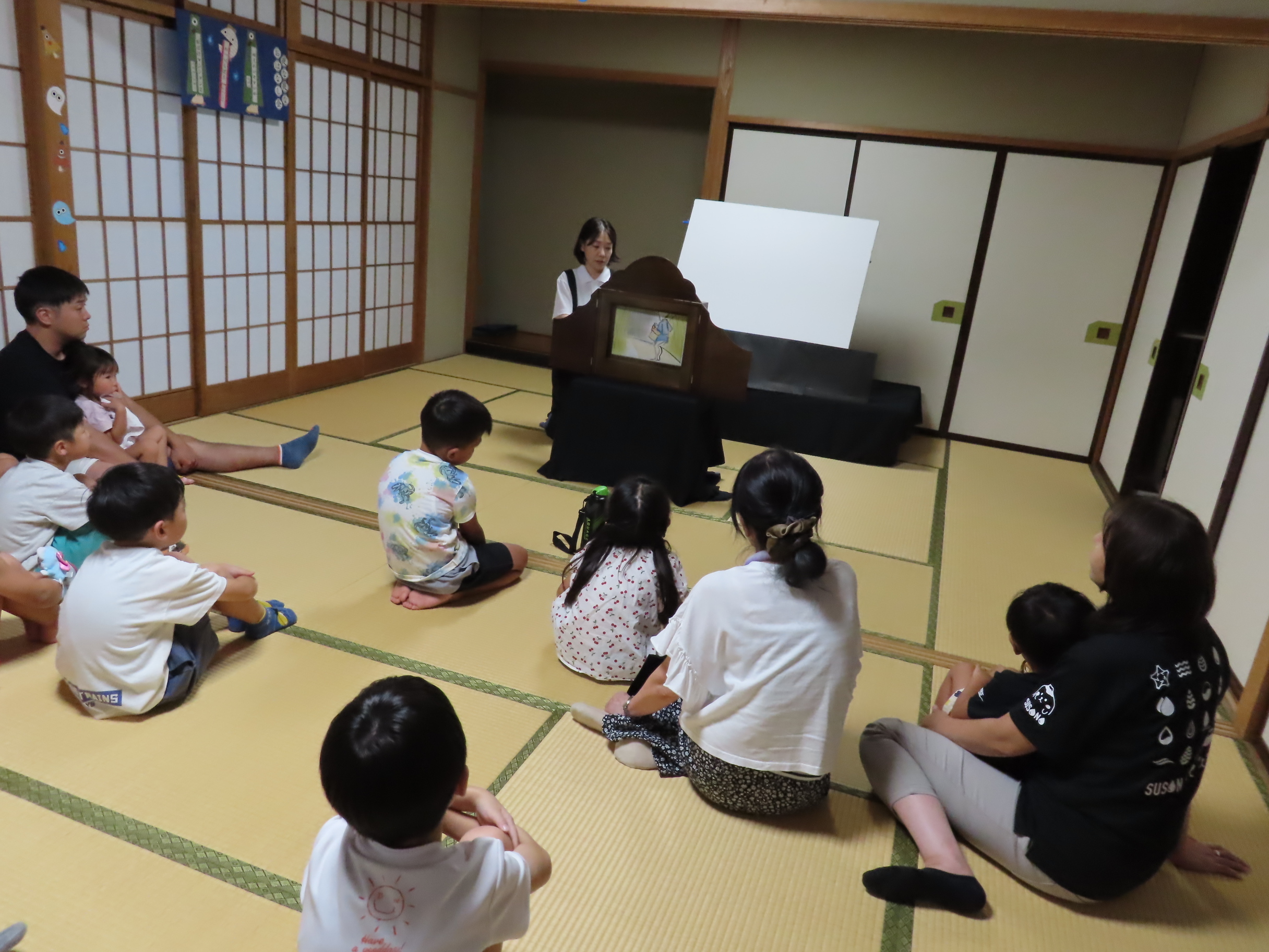 畳の部屋で座って紙芝居を見ている子どもたちと語り手