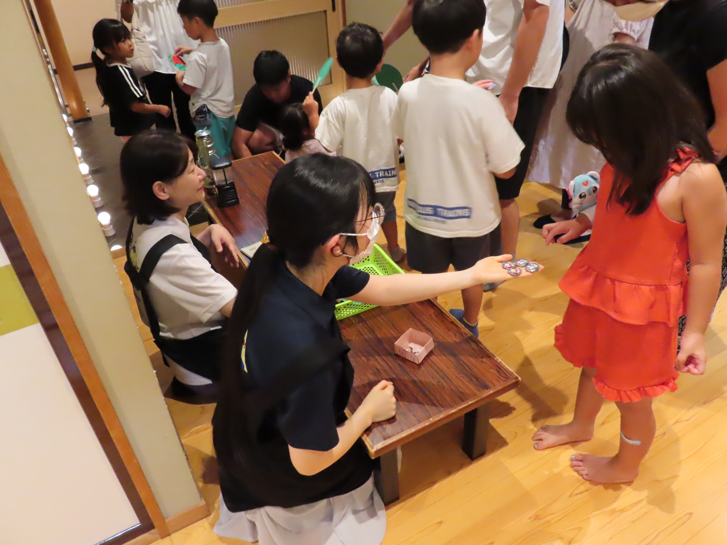 室内で複数の子どもたちが集まって職員が手渡しで缶バッジや手作りうちわを差し出している様子