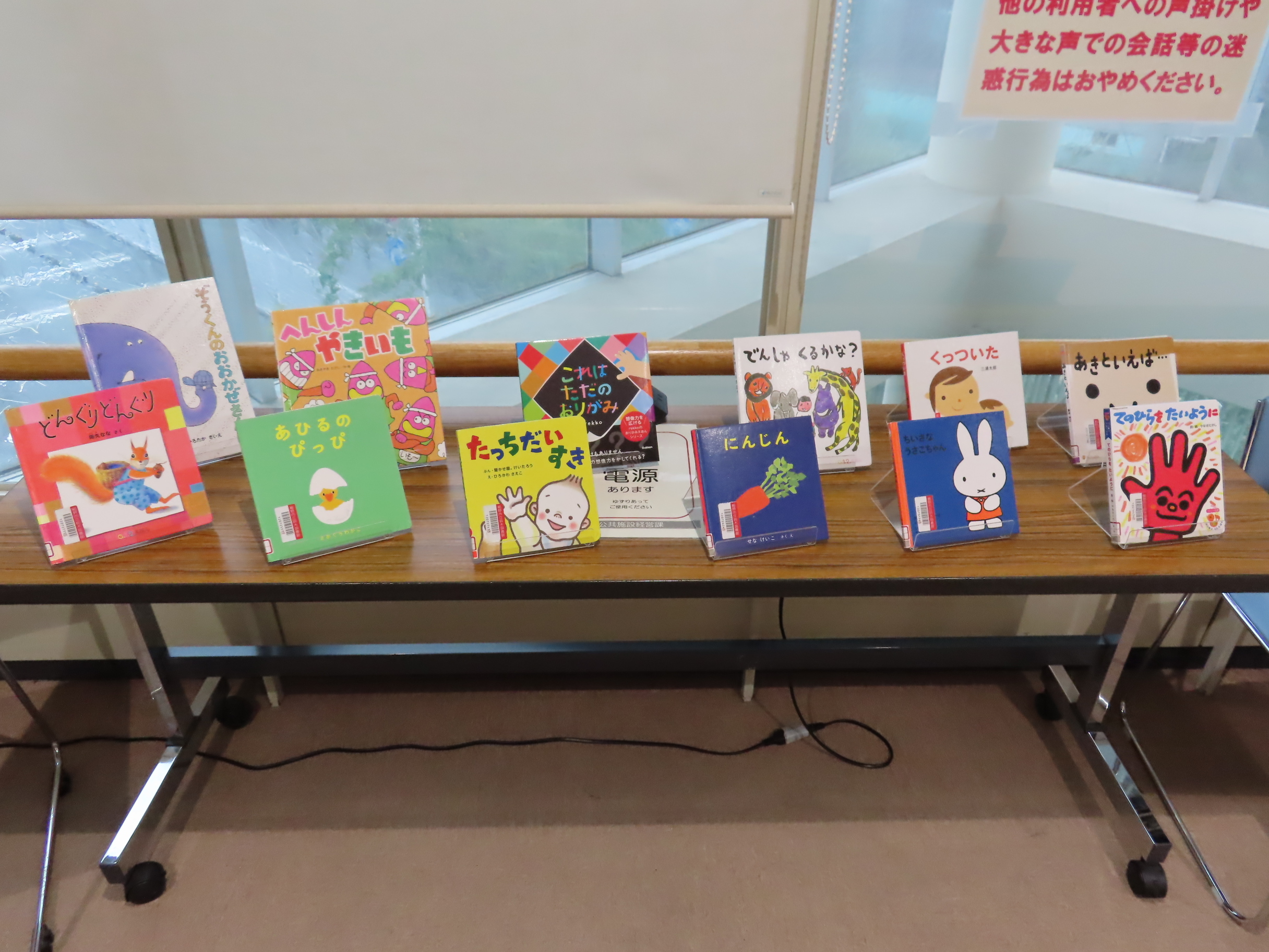 窓際の長いテーブルの上に並べられたカラフルな絵本が複数冊表紙が見えるように展示されている