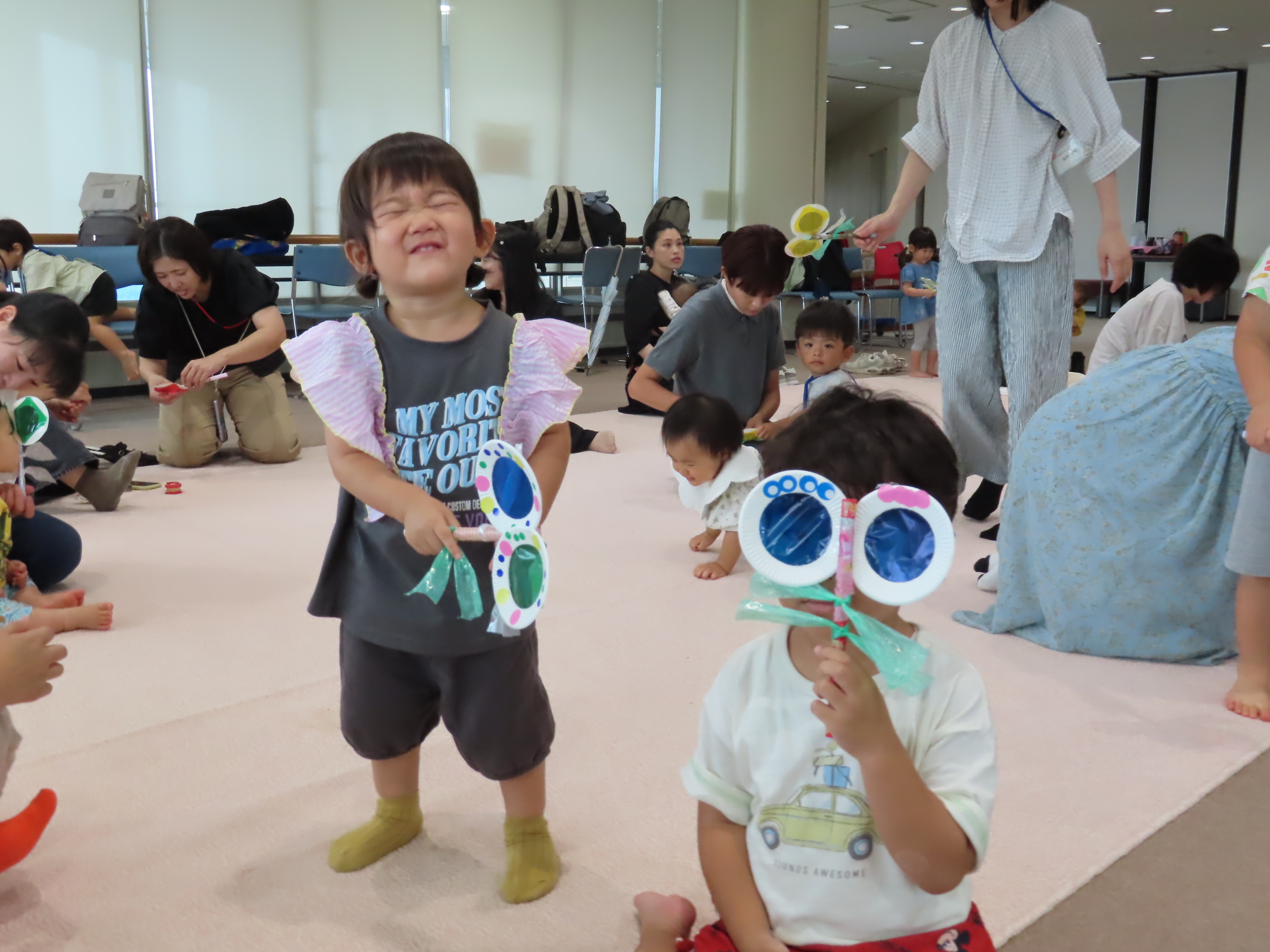 屋内の広い部屋で色とりどりの手作り眼鏡を持って遊ぶ幼児たち