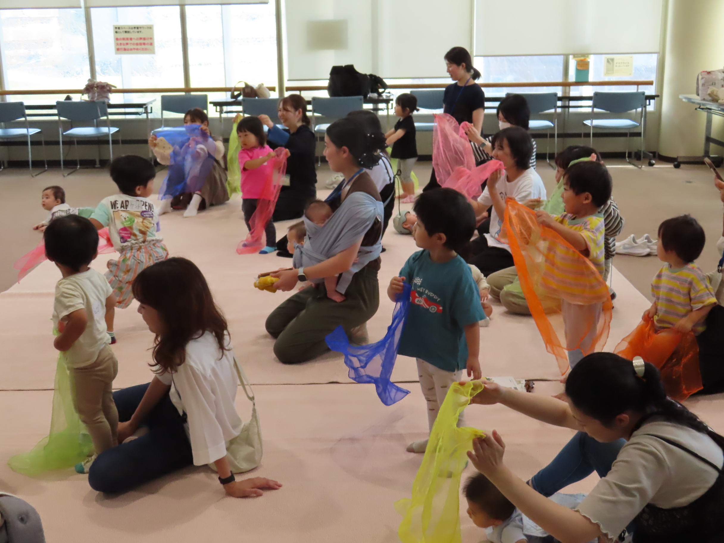 室内で幼児と保護者がカラフルな布を持って座ったり立ったりして遊んでいる講座の様子
