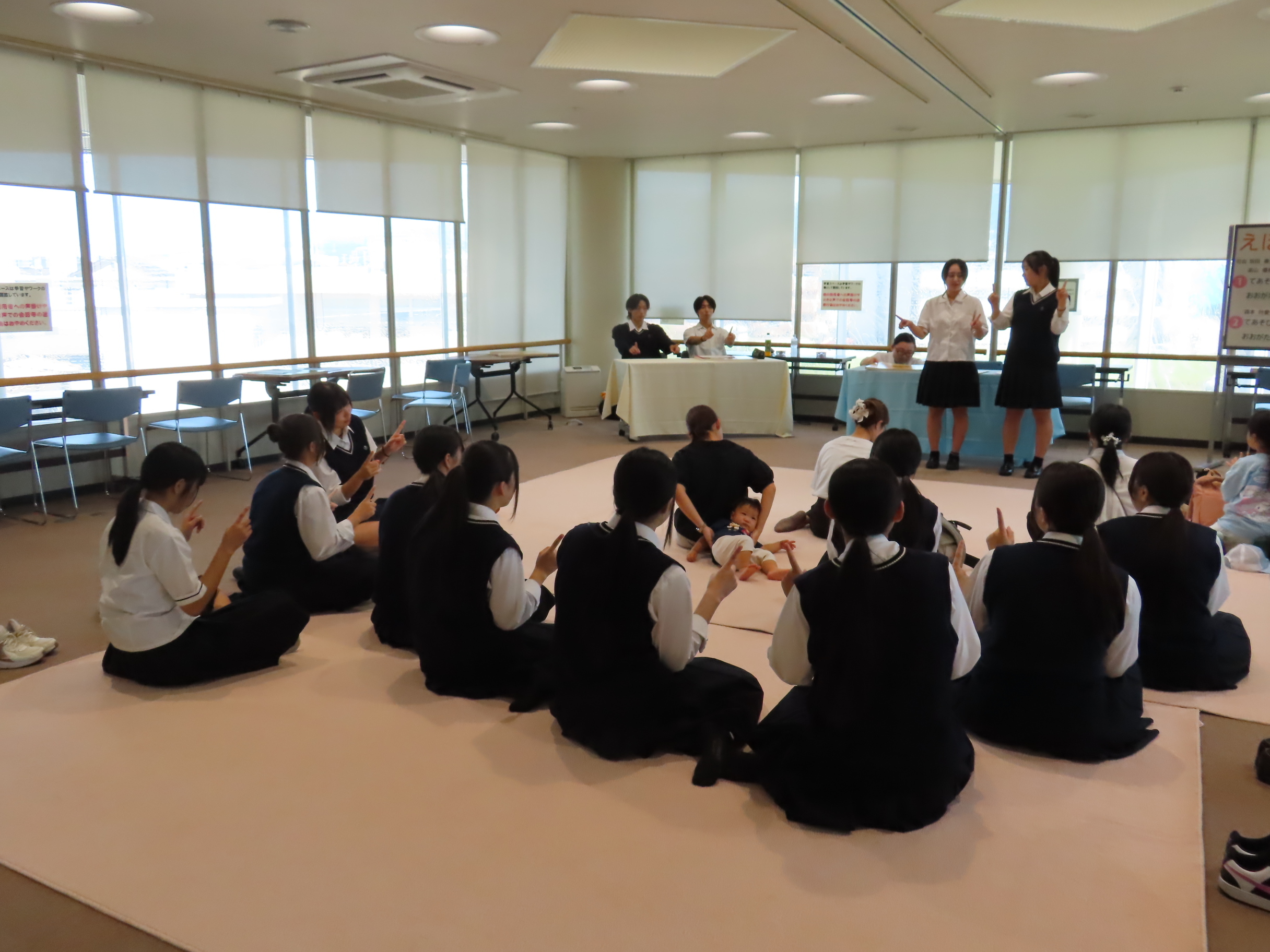 展示室の床に座って発表を聞く制服姿の生徒や幼児たちと前に立つ数人の発表者がいる風景