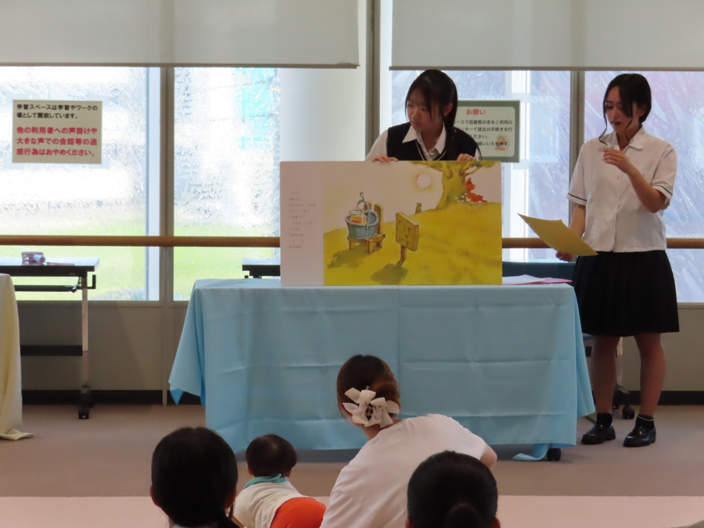 子どもたちが前方のテーブルに置かれた大型絵本を見て二人の発表者が立っている室内の光景
