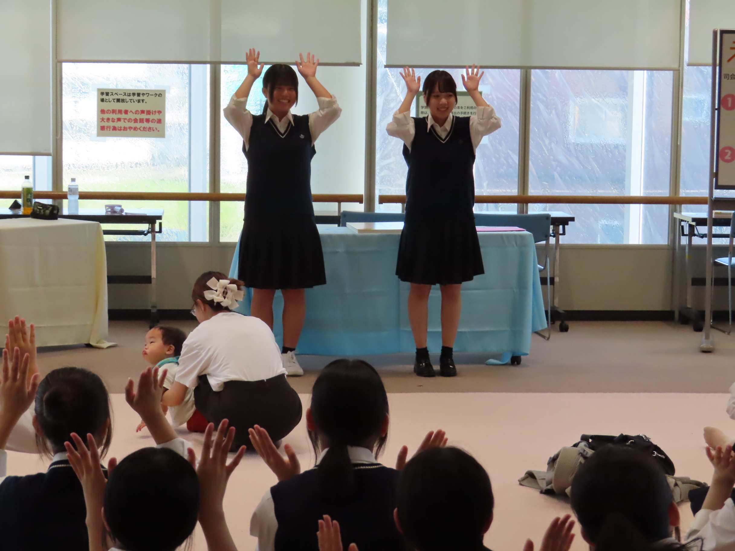 展示室の前で制服を着た二人の生徒が両手を上げて立ち床に座った子どもたちがそれを見ている光景
