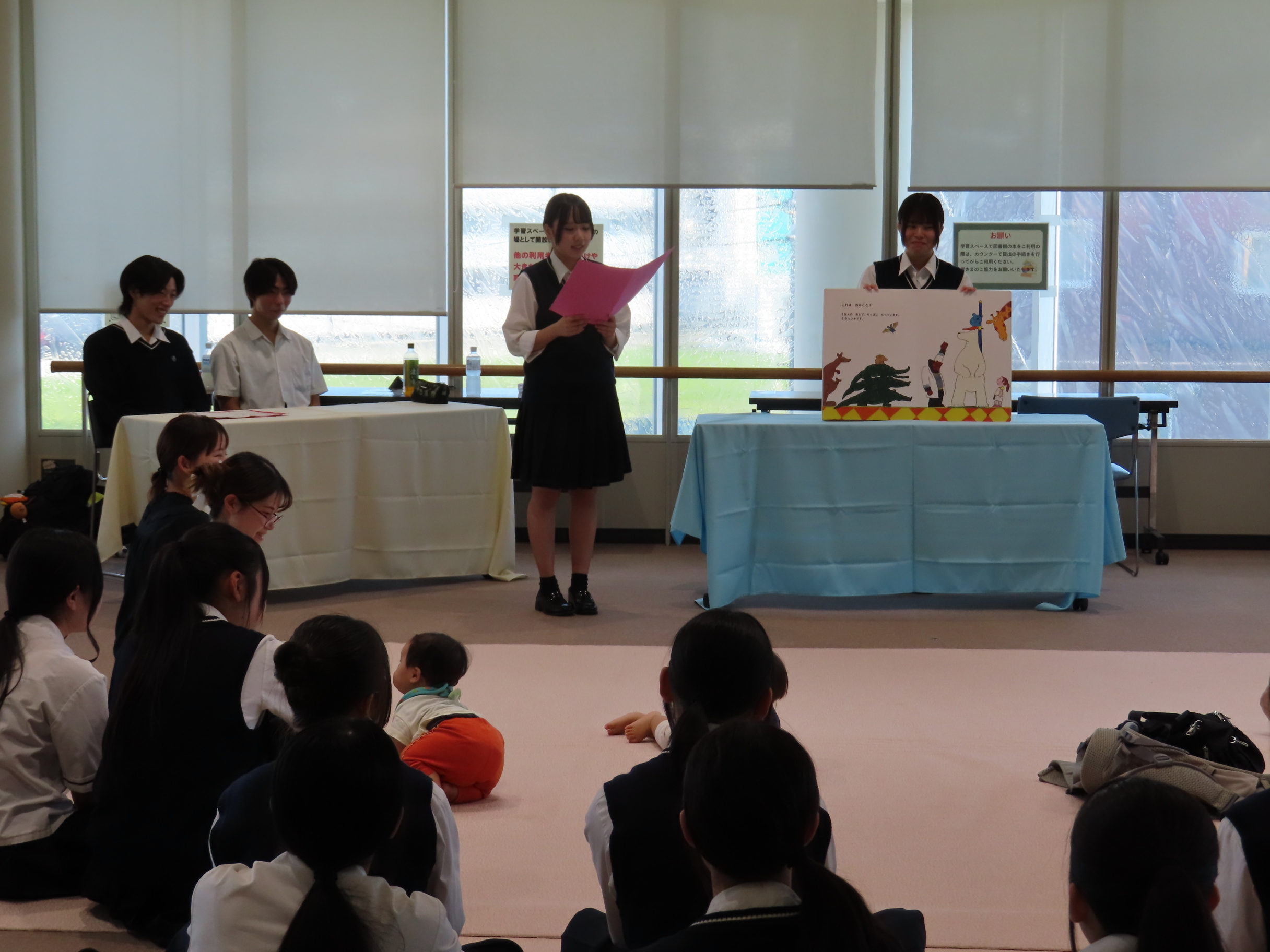 展示室で床に座る乳幼児と保護者たちが前方の机の上で大型絵本の読み聞かせする生徒を見ている様子