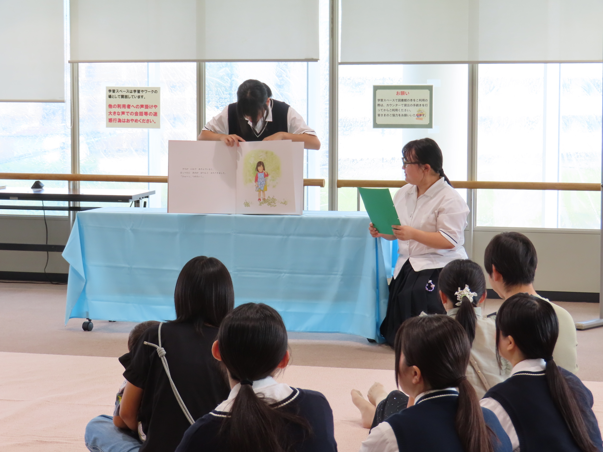 展示室で座る子どもたちが大型絵本の読み聞かせを見ている光景