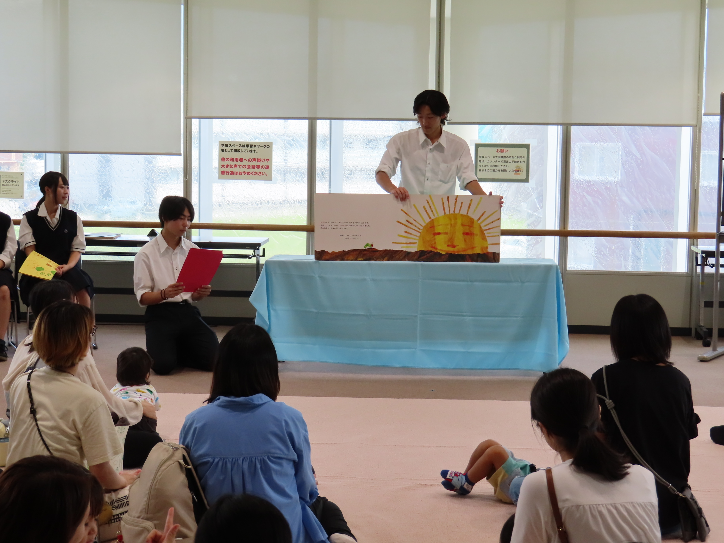 テーブルの上の大型絵本を使って室内で子どもと保護者の前で読み聞かせをする高校生