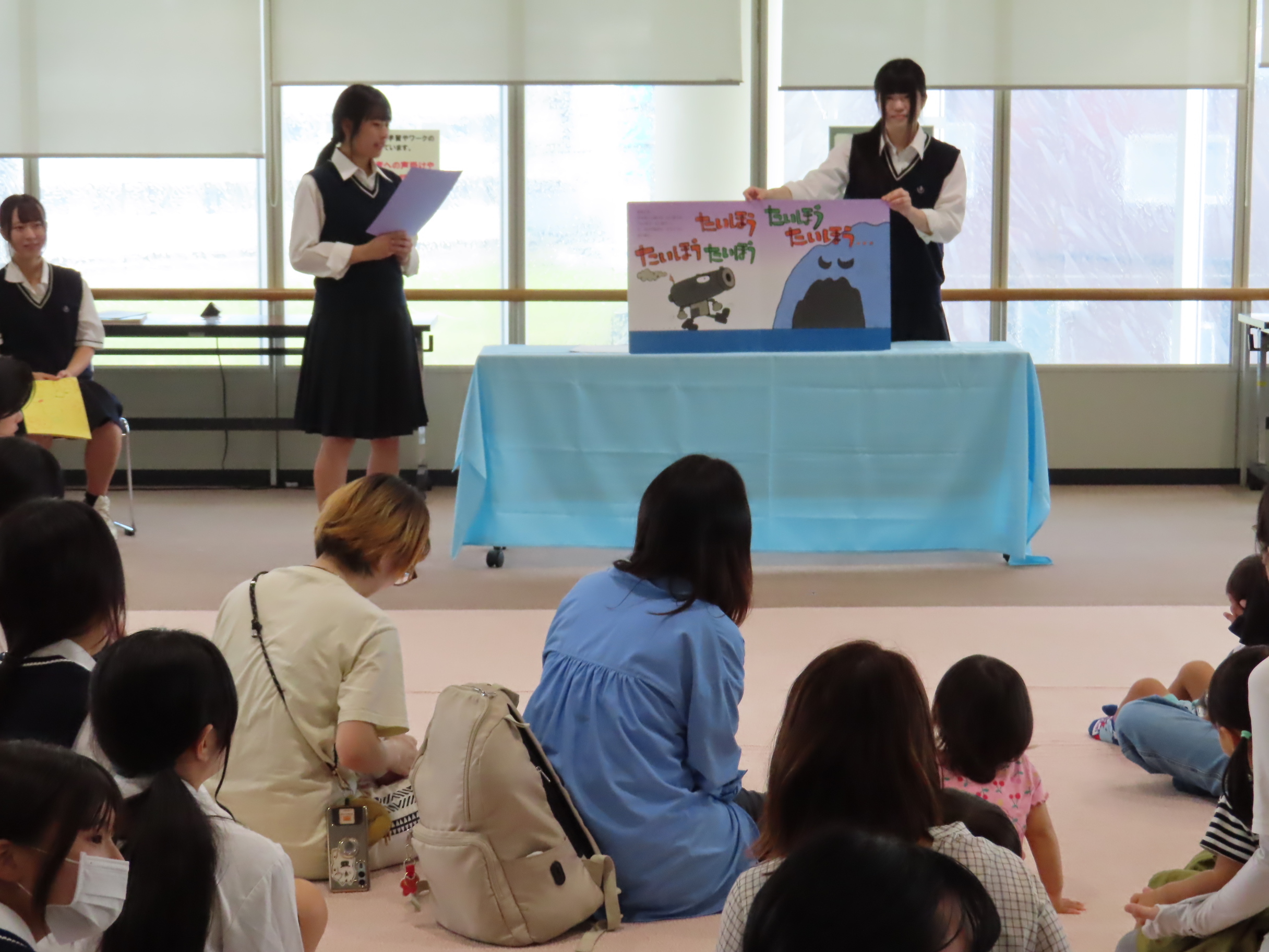 展示室の前で立った制服姿の二人が大型絵本を使って発表していて床に座った子どもや保護者たちがそれを見ている光景
