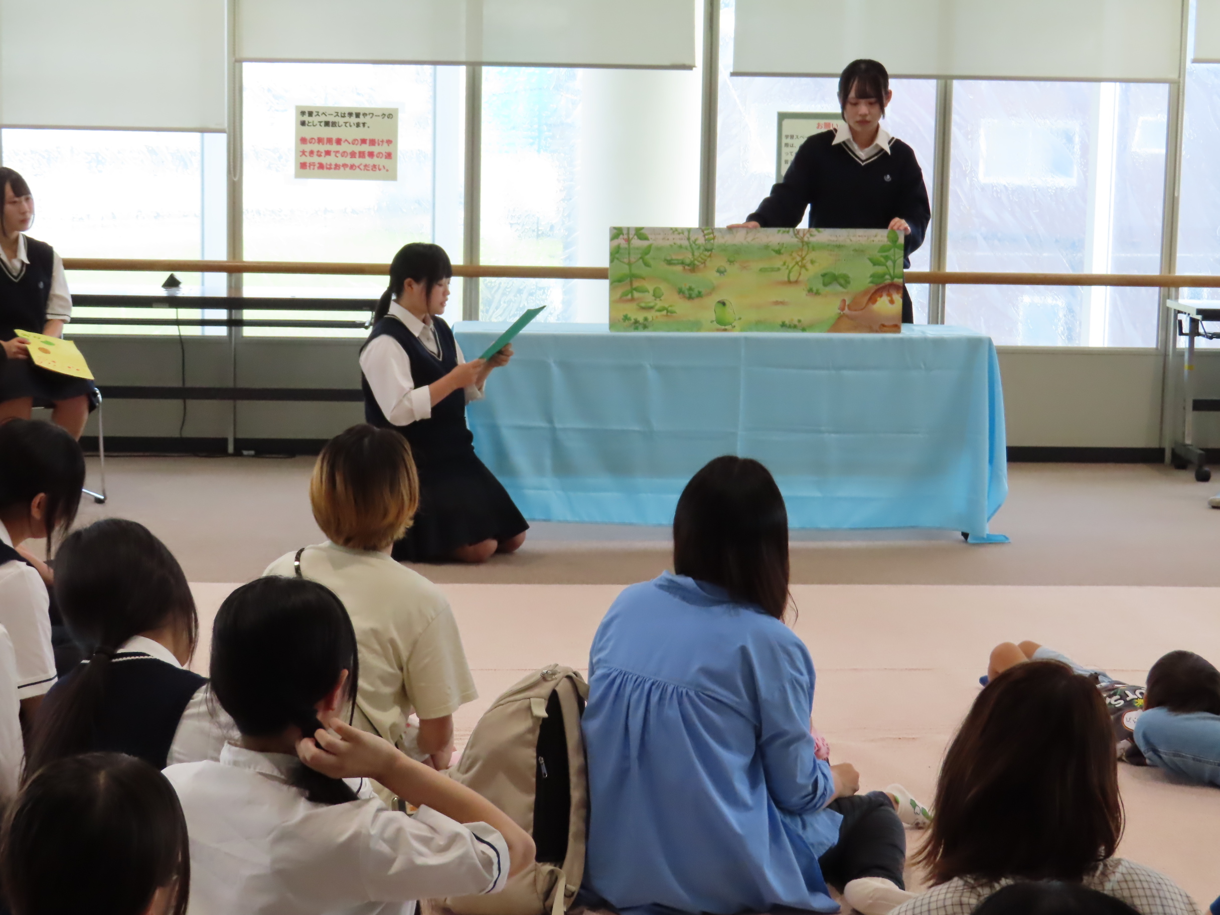 数人の学生や子どもたちが床に座って前方の読み聞かせを見ている様子