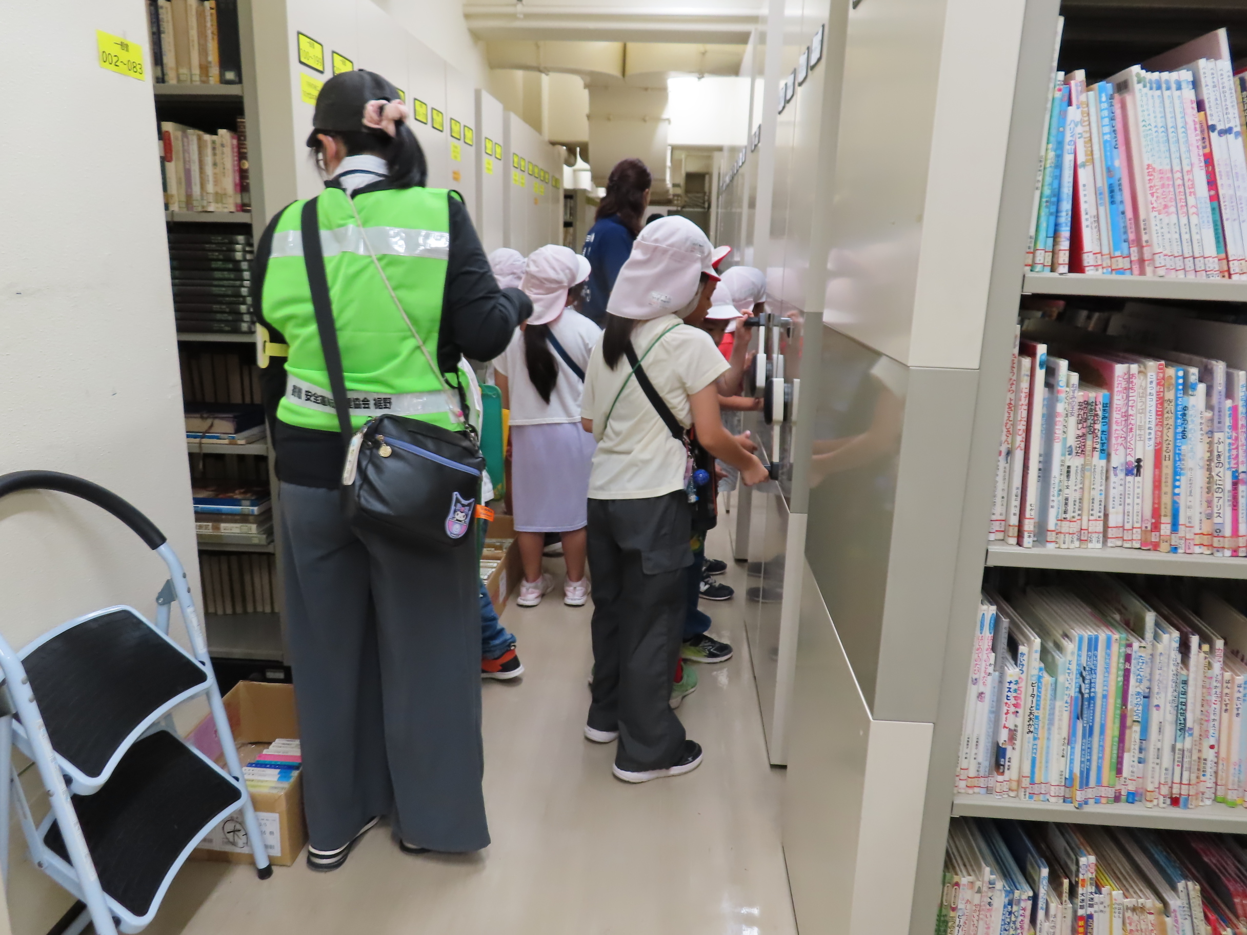 図書館の書架の間で蛍光緑のベストを着た大人と白い帽子をかぶった子どもたちが列になって本を探している様子