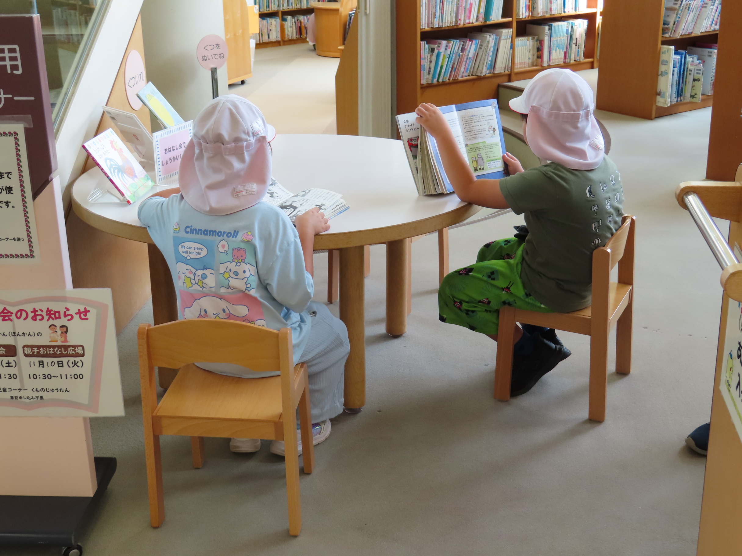 図書館の児童コーナーで帽子をかぶった二人の子どもが丸テーブルで本を読む