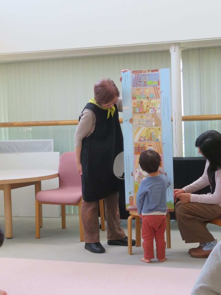 子供が大人に向かって絵本を見ている場面で、周囲には椅子とテーブルがある室内の様子。