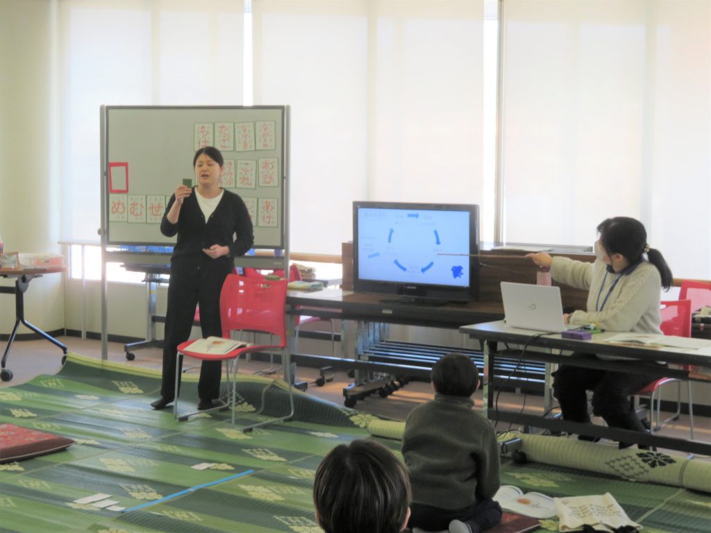 教室でプレゼンテーションを行う人物とコンピュータを操作する別の人物がいる様子が映っている画像。