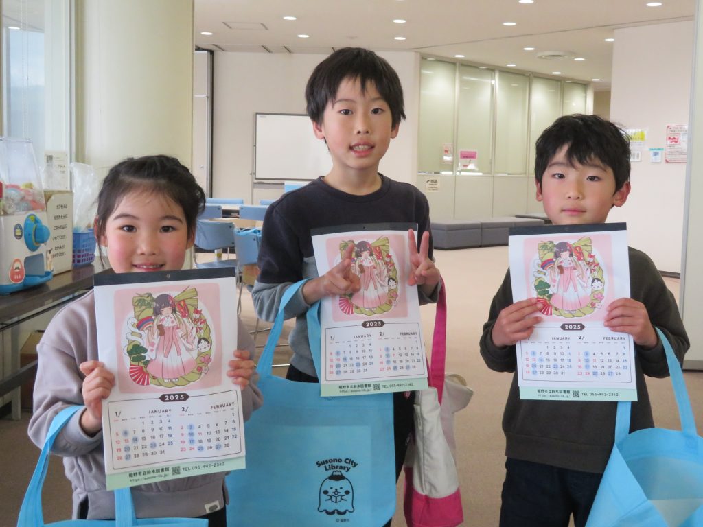 3人の子供がカラフルなバッグを持ち、賞状を掲げている写真。