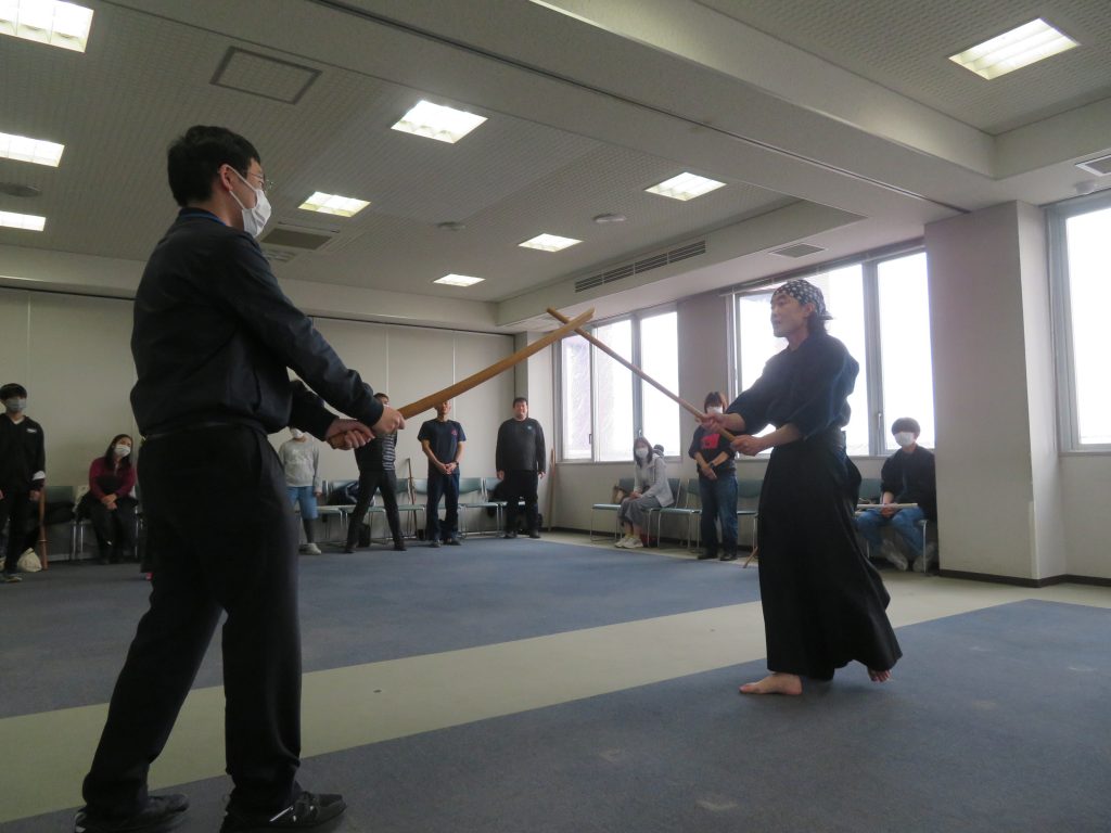 二人の武道家が道場で木刀を持って対峙している様子